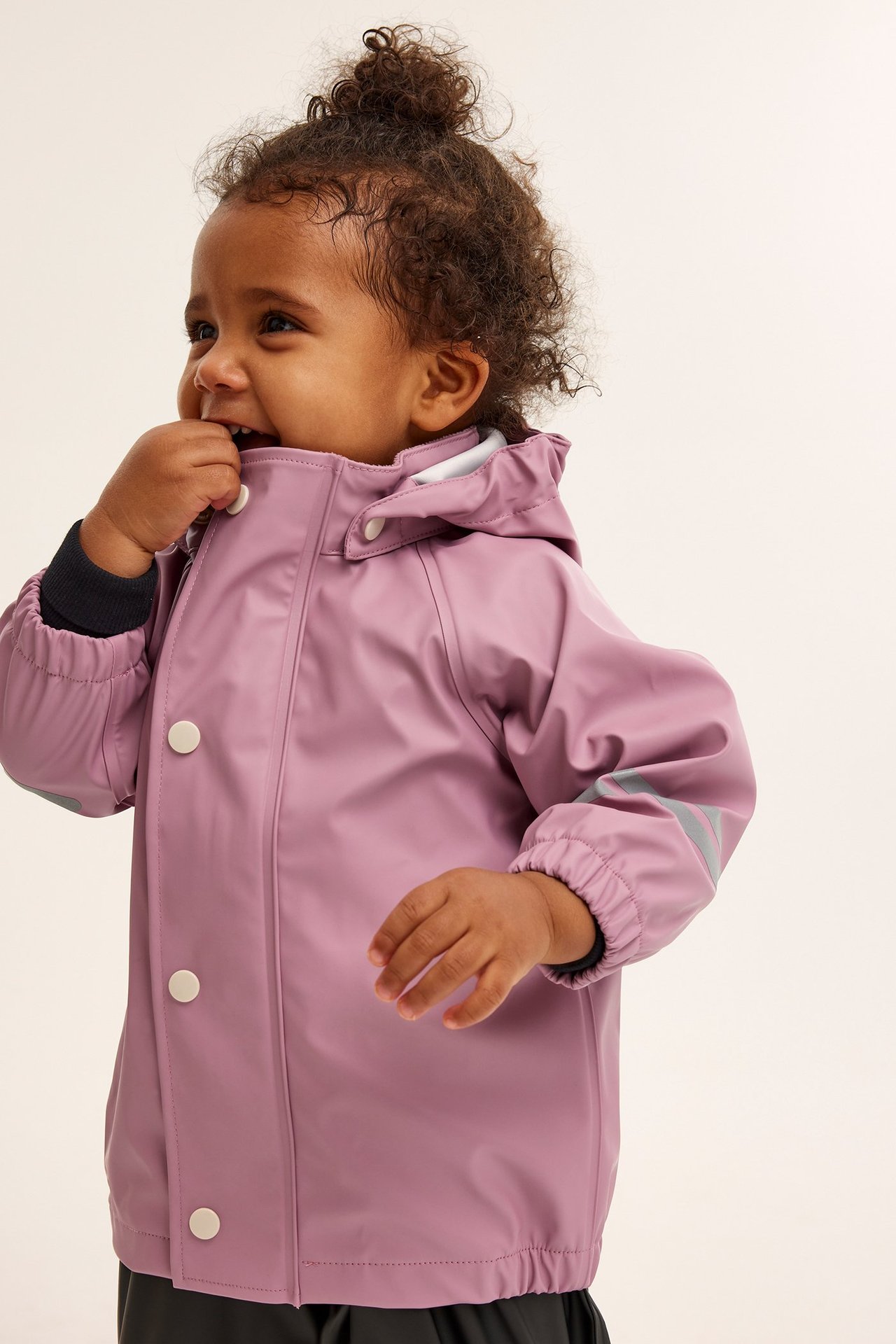 Sidebilde av en baby i rosa regnjakke med hvite knapper og refleksstriper på ermet.