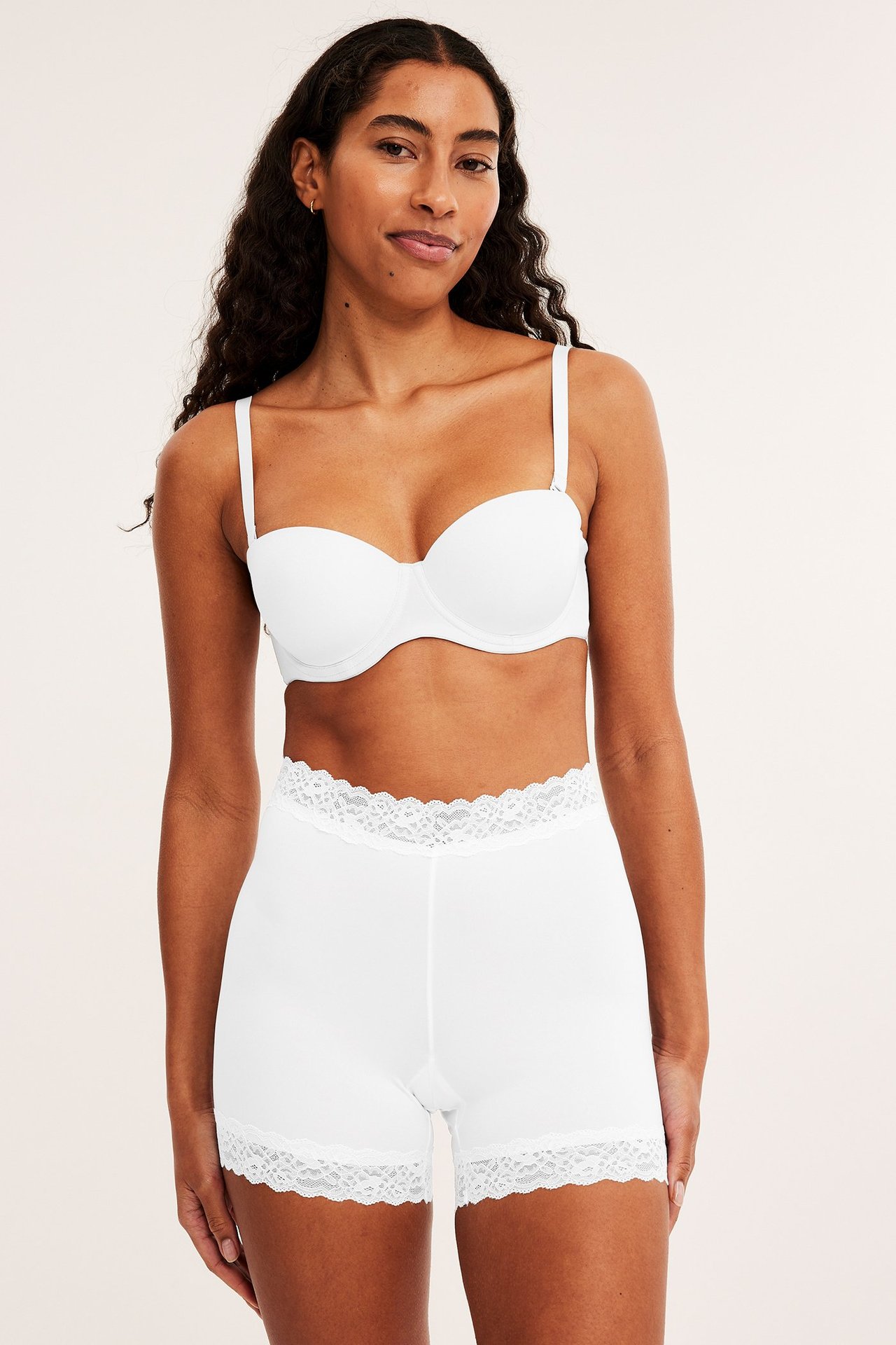 Frontbilde av en kvinne i hvite mikroshorts med blondekanter og en hvit stroppeløs BH.