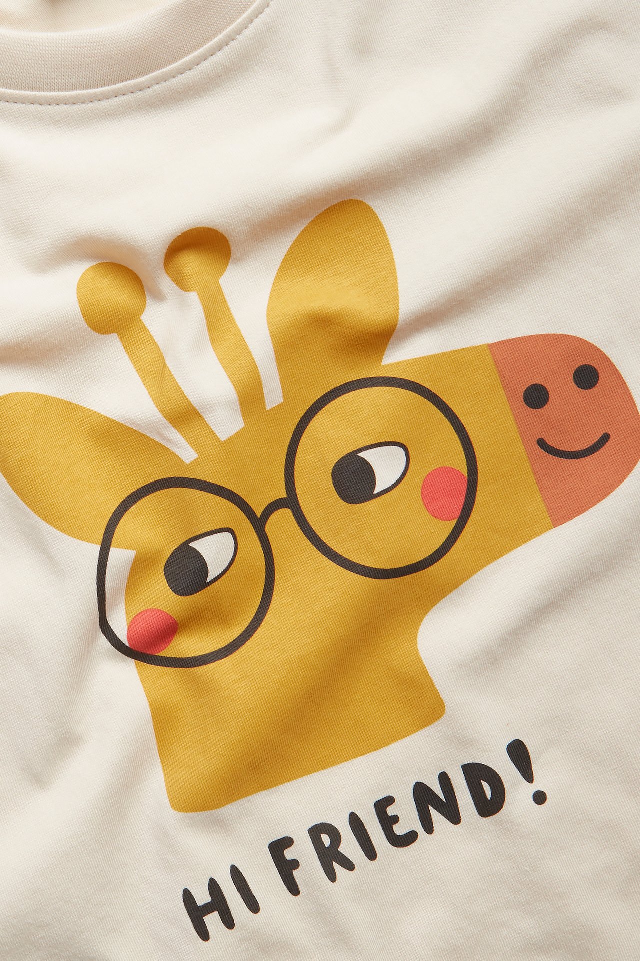 Nærbilde av en beige bomulls-T-skjorte for barn med trykk av en sjiraff med briller og 'HI FRIEND!'.