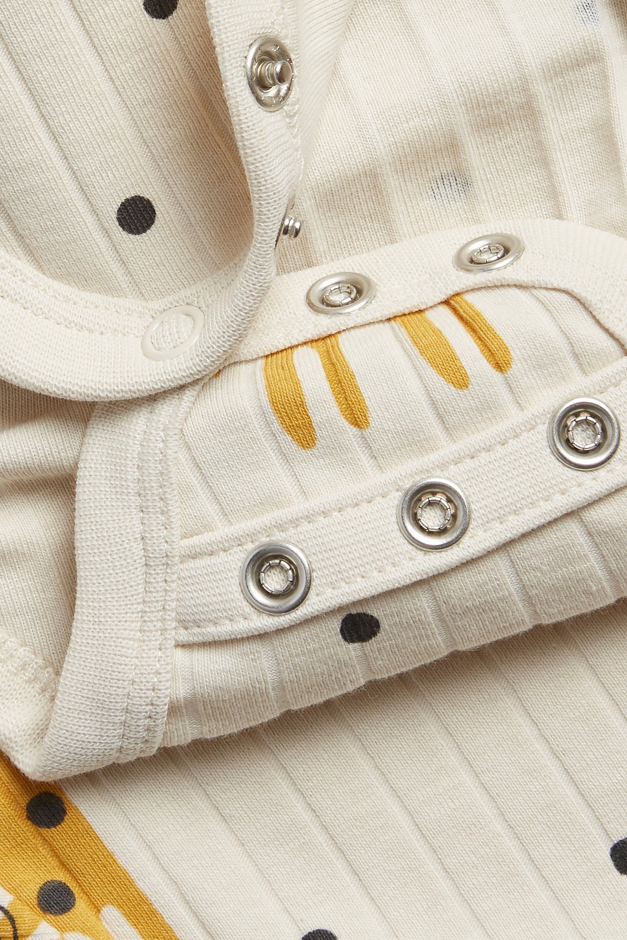 Närbild av en beige ribbad babybody med svarta prickar och gult djurmönster, med tryckknappar.