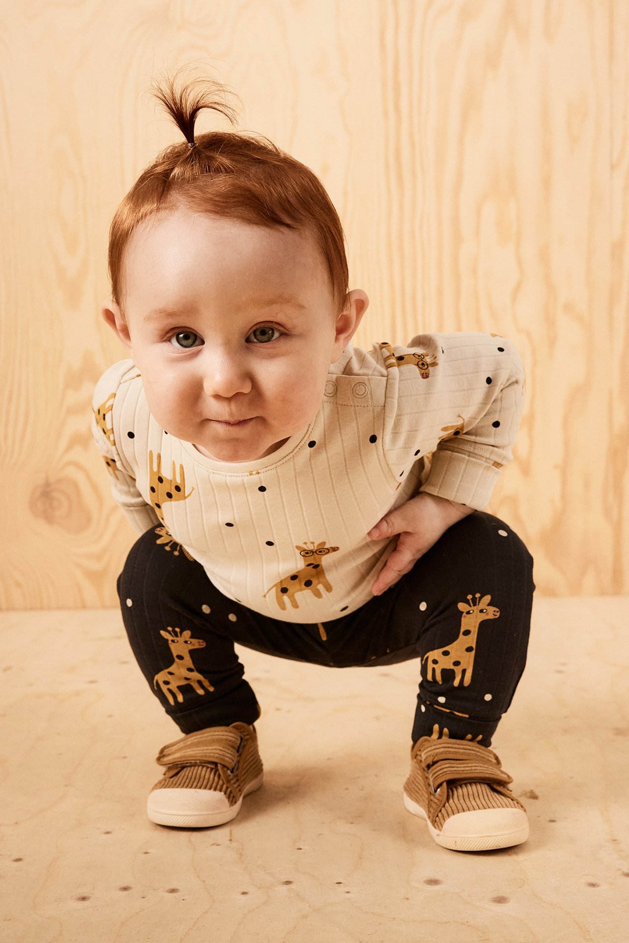 Framifrån, baby i beige ribbad body med giraffmönster, mörka giraffmönstrade leggings och bruna skor.