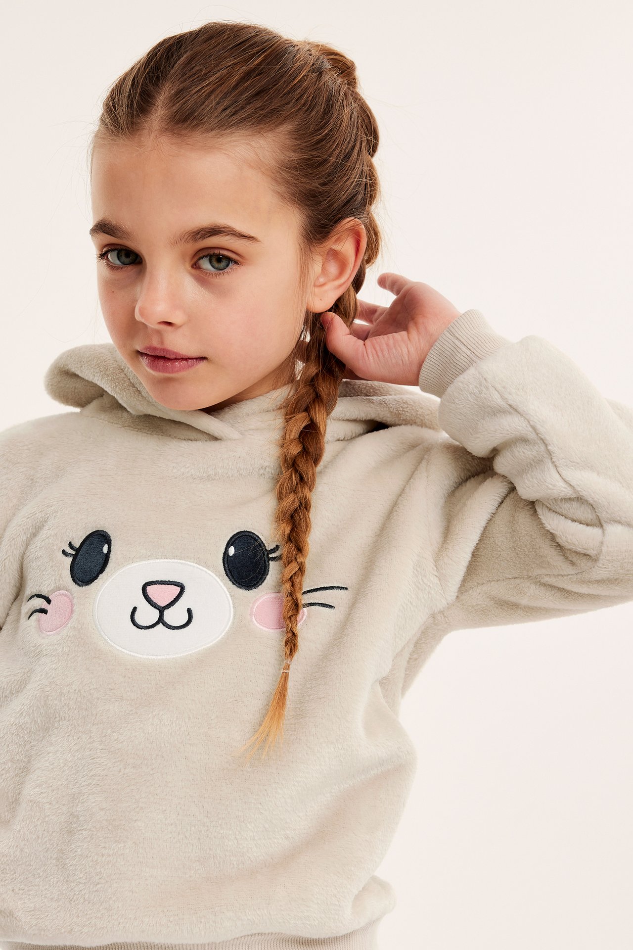 Nærbilde forfra av et barn i en beige lodden fleecehettegenser med brodert katteansikt.