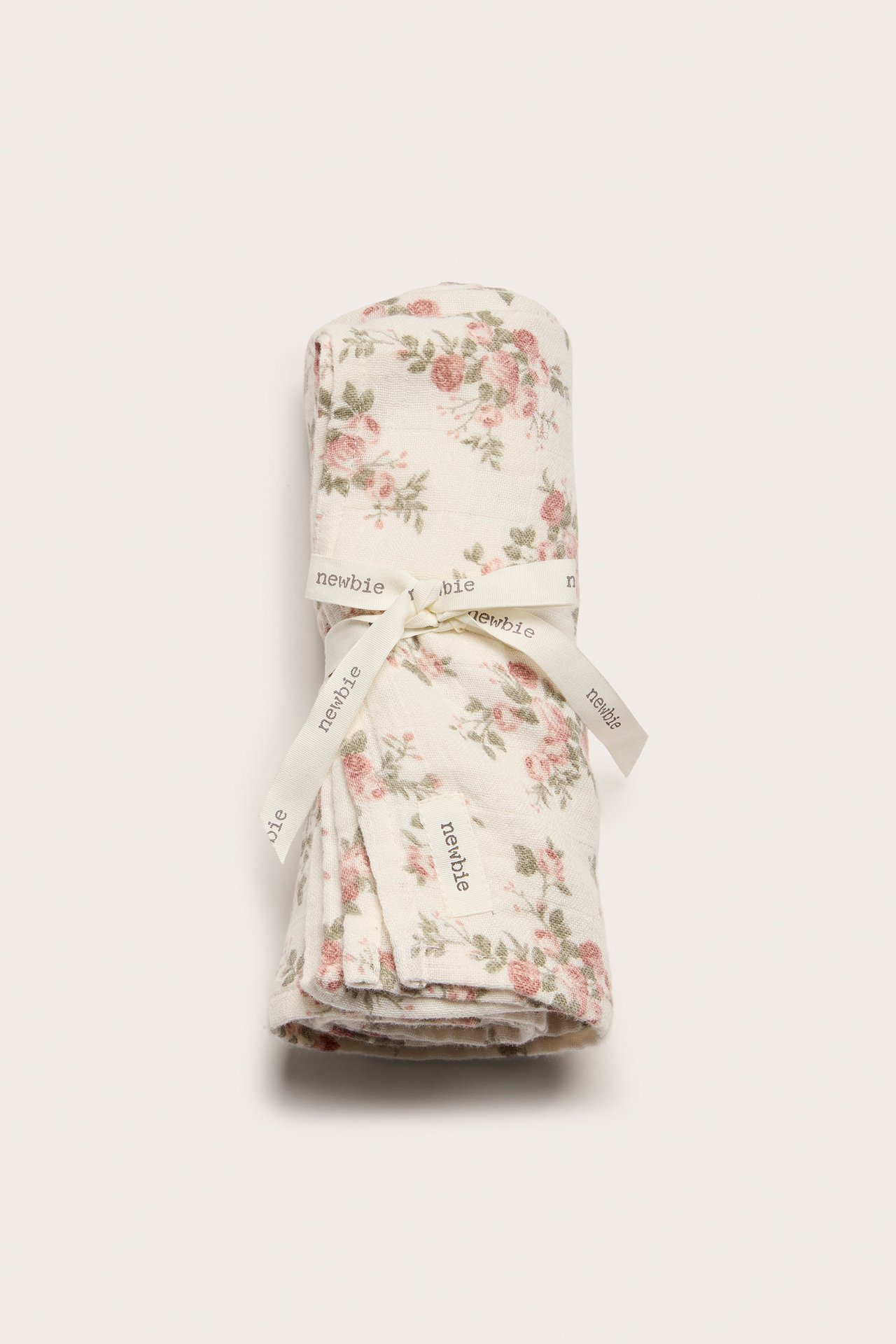 Framifrån: en offwhite bomullsmuslinfilt med rosa blommönster, rullad och knuten med ett band.