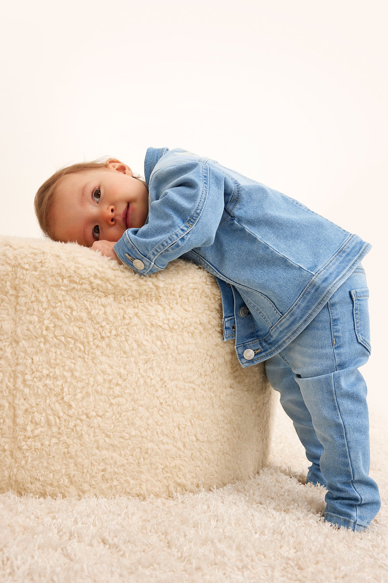 Framifrån: Baby i ljusblå denimjeggings och matchande jeansjacka, lutande mot en gräddvit puff.