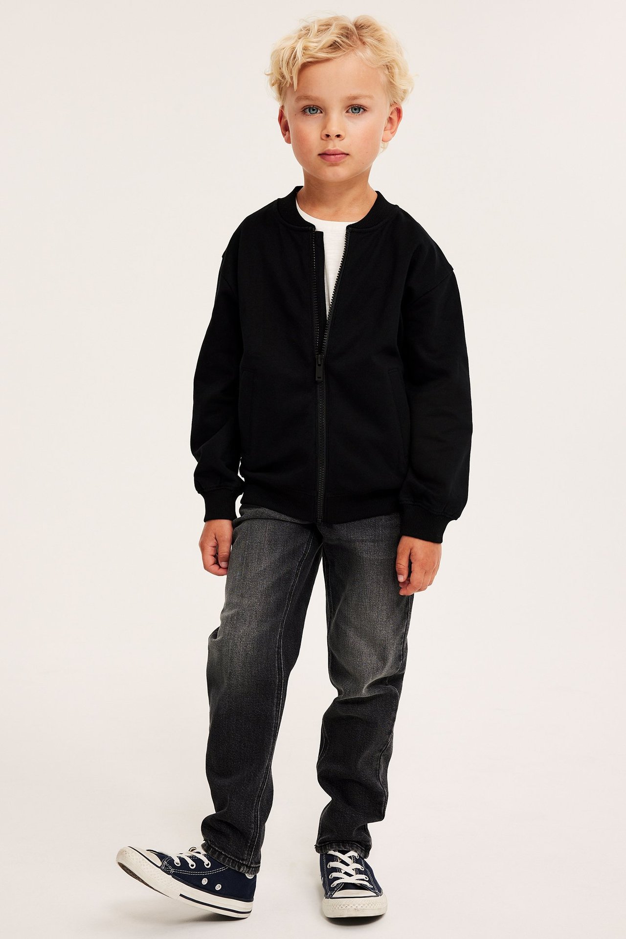 Framifrån, ett barn i svart bomullskofta med dragkedja, vit t-shirt, mörkgrå jeans och marinblå sneakers.