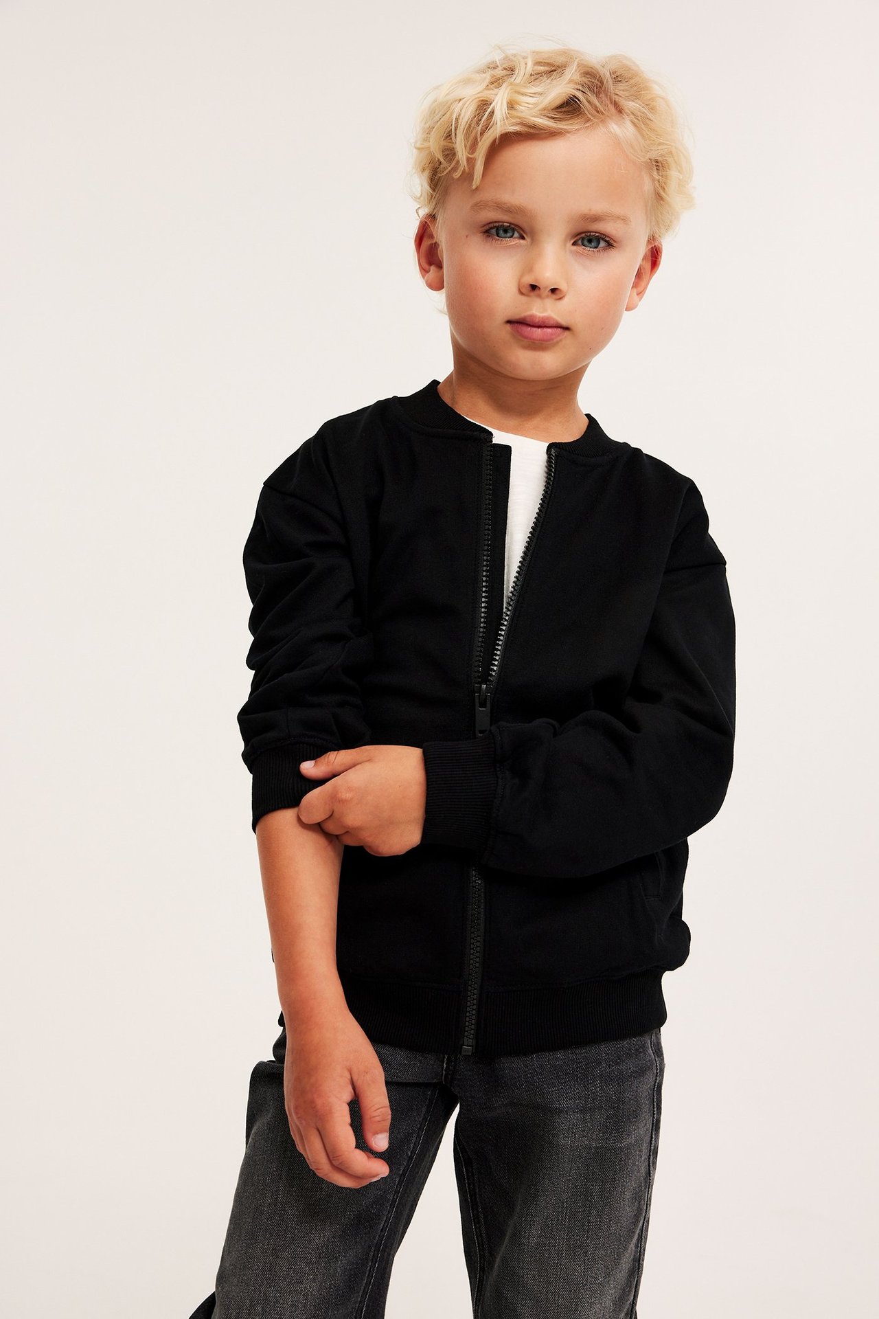 Widok z przodu dziecka w czarnym bawełnianym rozpinanym swetrze, białym t-shircie i ciemnoszarych dżinsach.