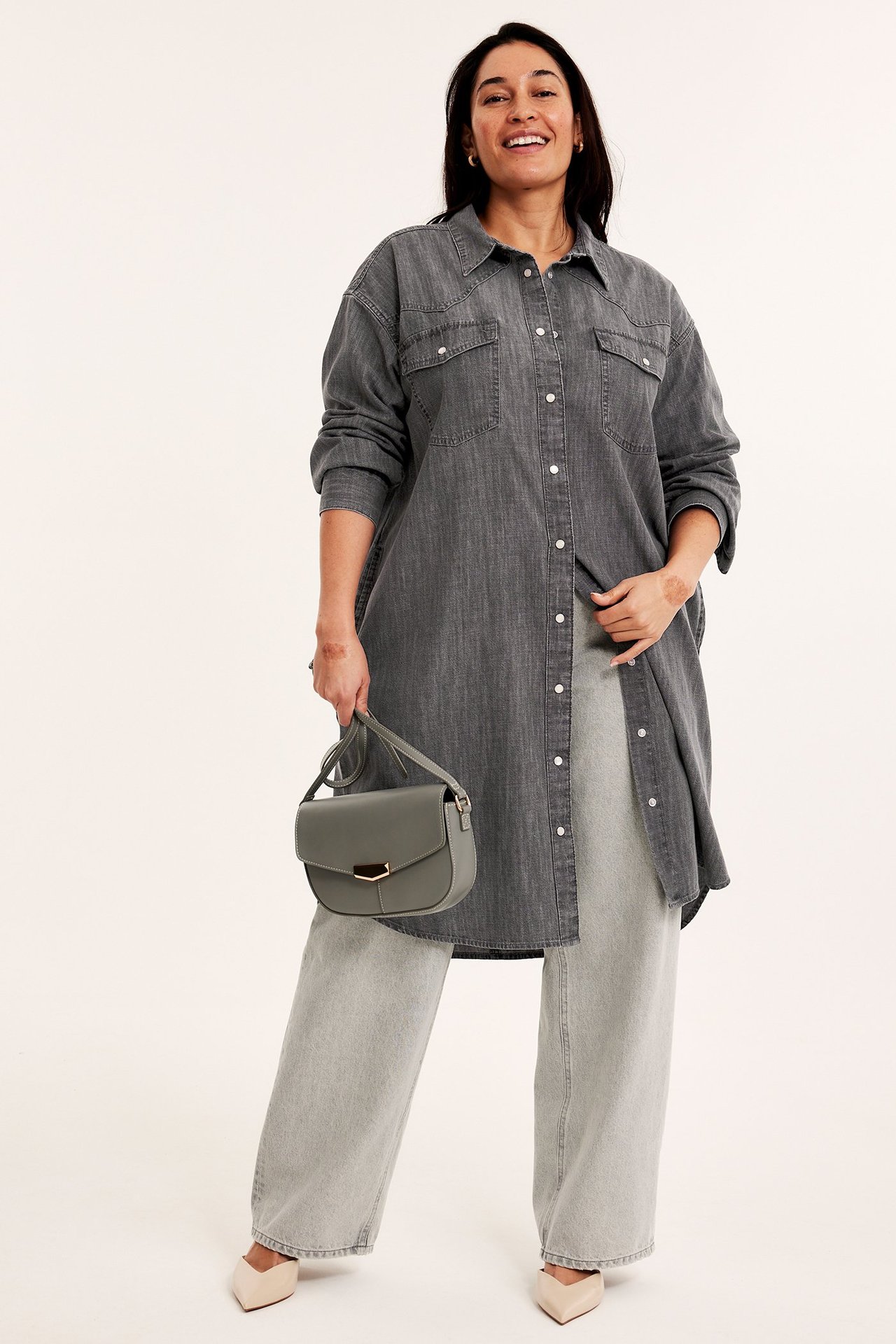 Widok z przodu: Kobieta w szarej dżinsowej sukience koszulowej, jasnoszarych szerokich spodniach, beżowych szpilkach, trzymająca szarą torebkę.