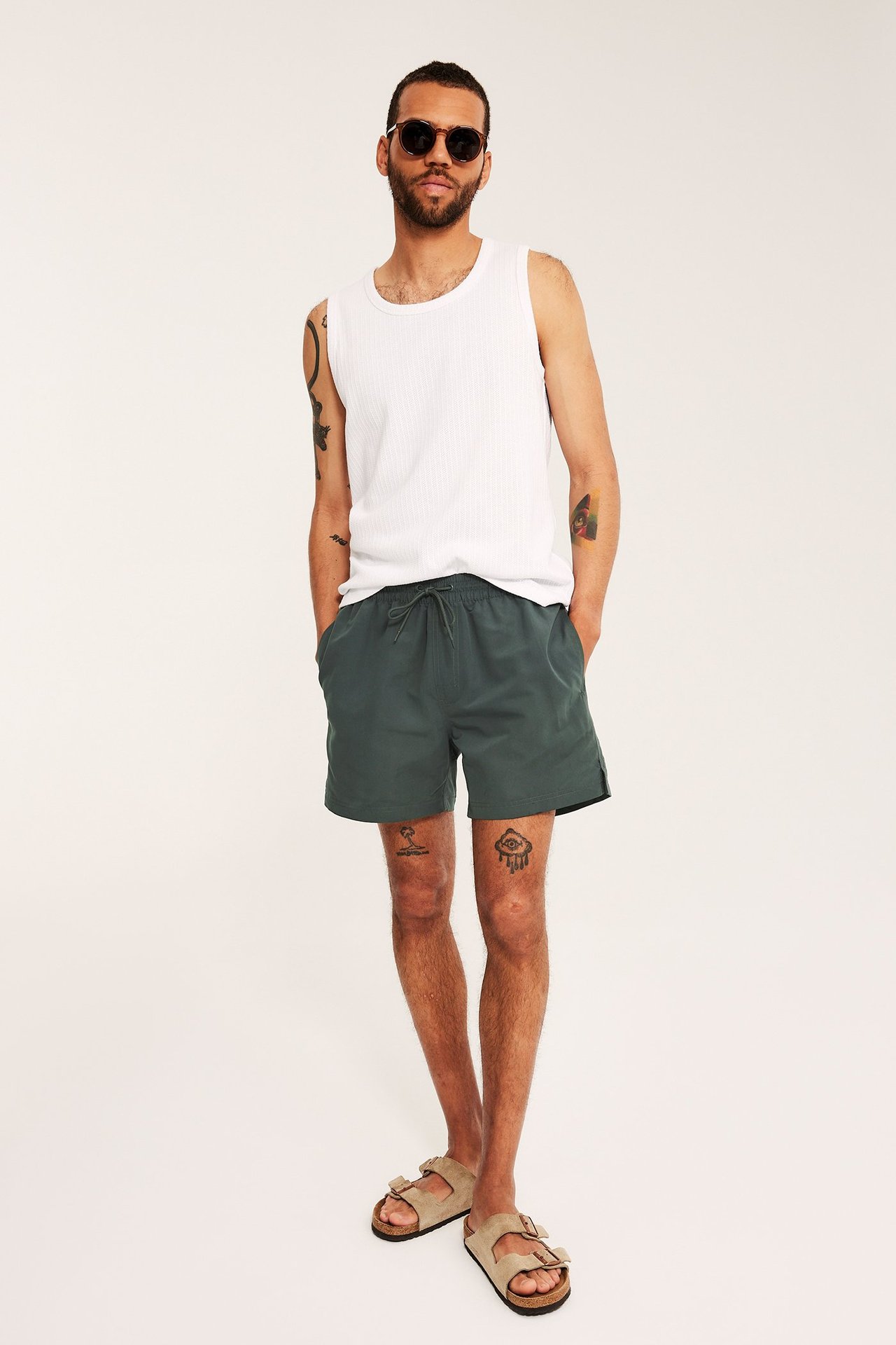 Edestä: Mies tummanvihreissä uimashortseissa, valkoisessa ribbitoppi, aurinkolasit, beiget sandaalit.