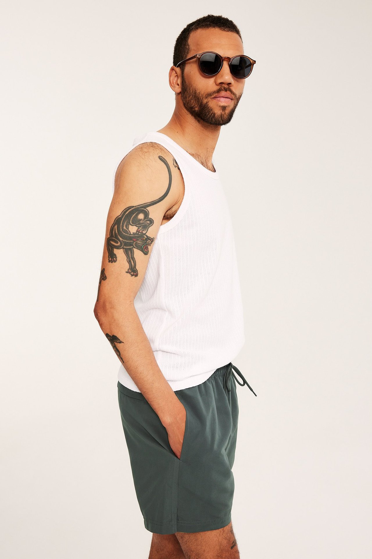 Widok z boku mężczyzny w ciemnozielonych kąpielówkach ze sznurkiem, białej koszulce i okrągłych okularach.