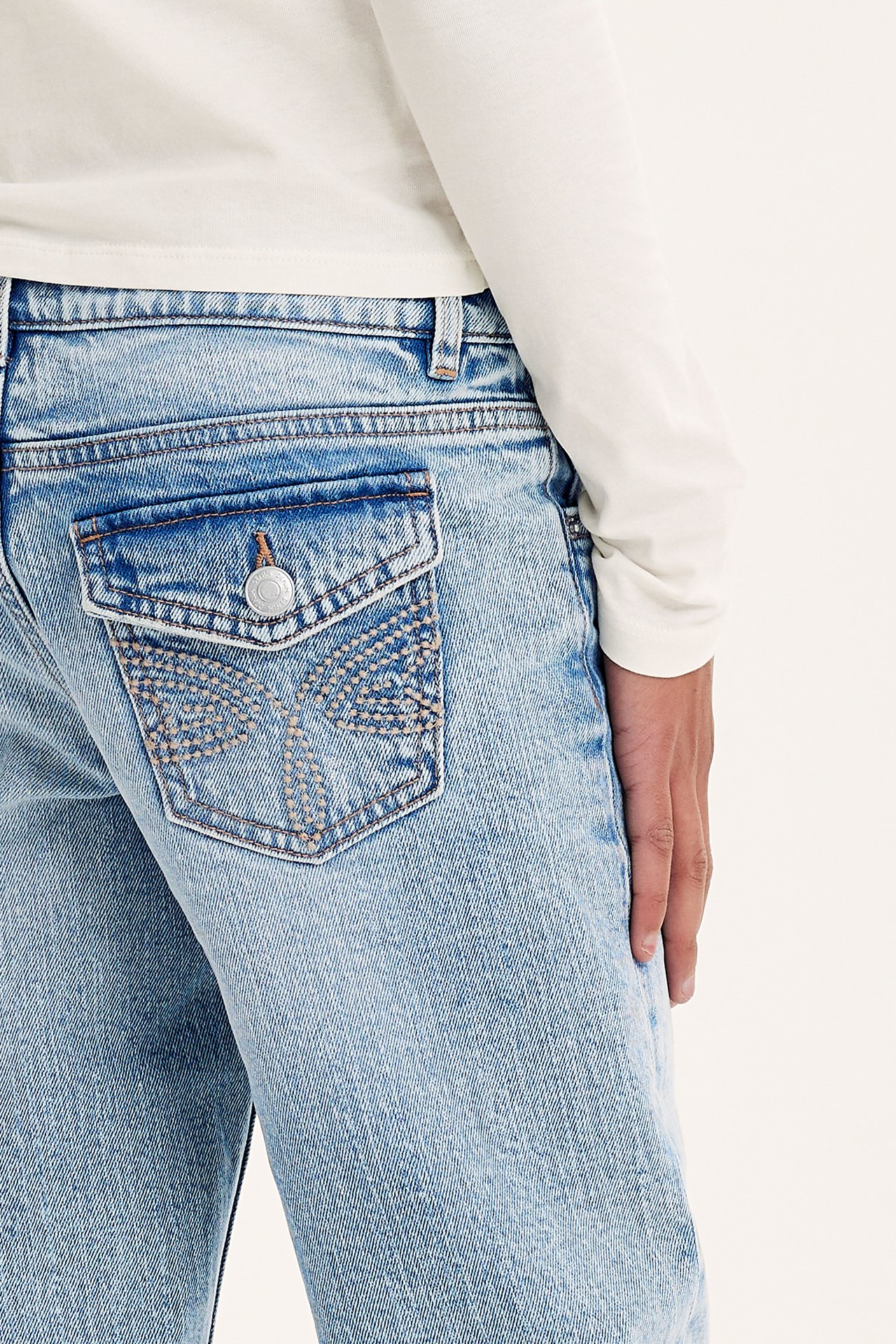 Närbild på barn i ljusblå bootcut-jeans med dekorativ bakficka med lock och vit långärmad topp.