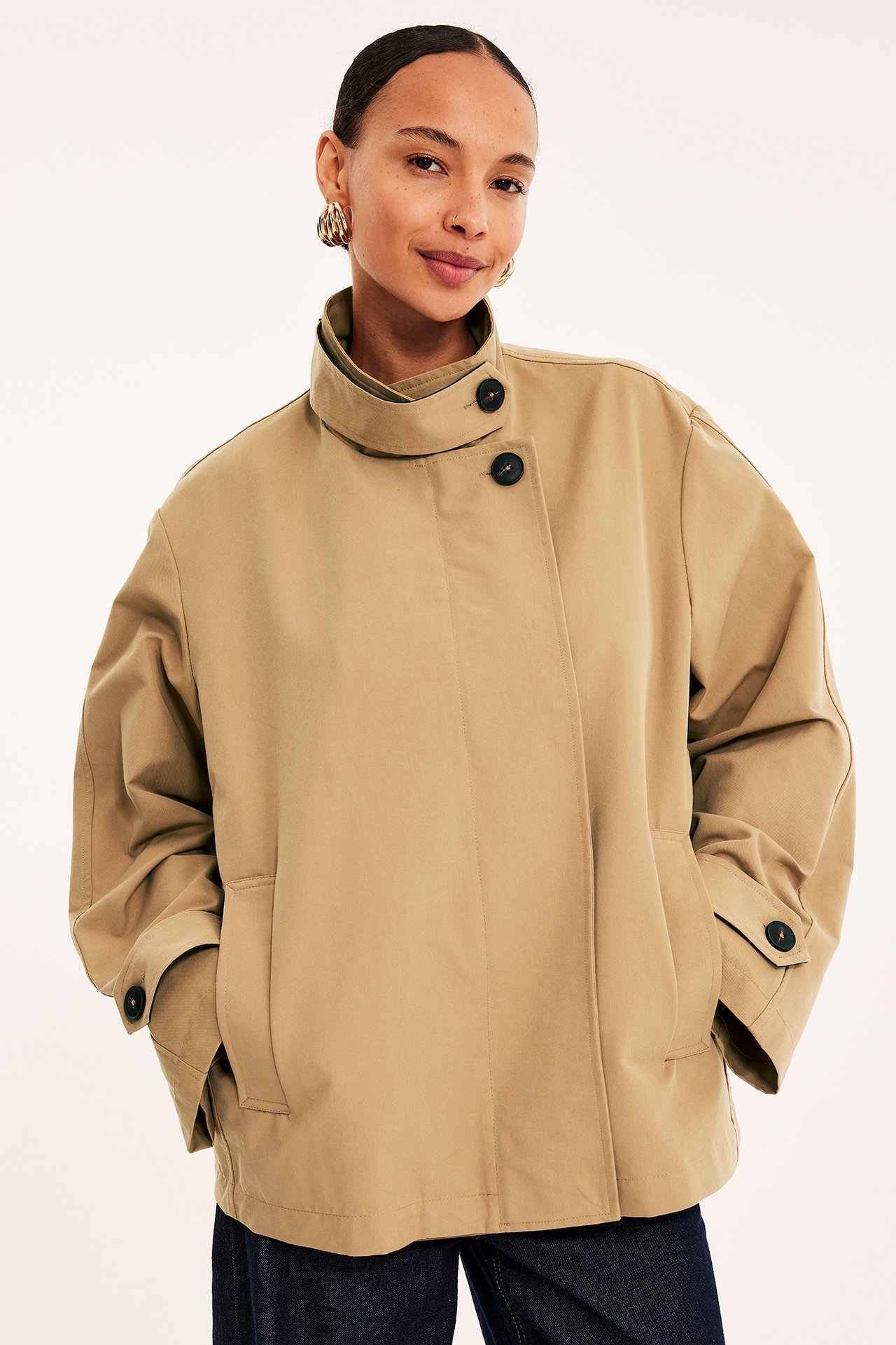 Forfra: kvinne i løs beige kort trenchcoat med asymmetriske knapper, mørke jeans og gulløredobber.