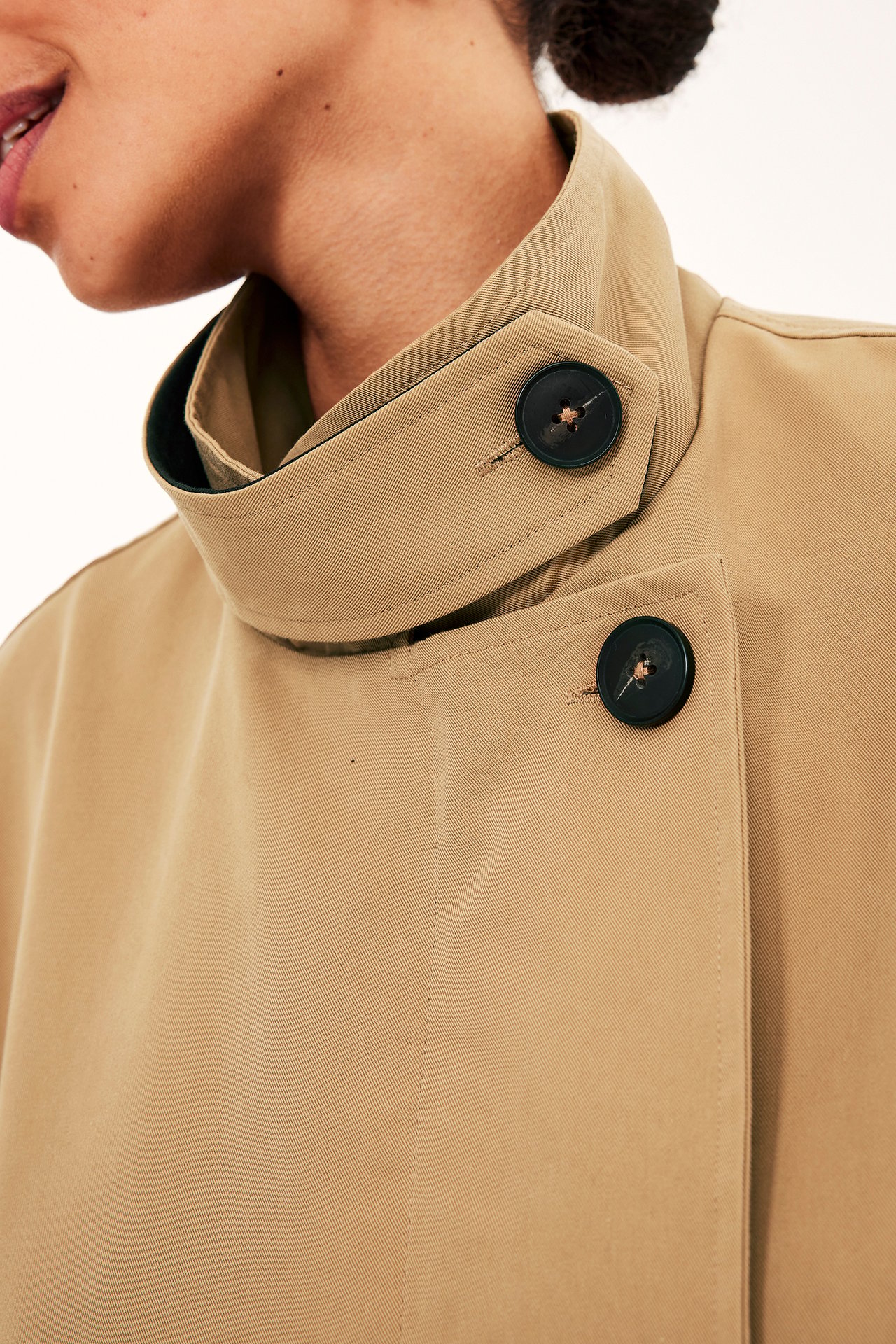 Nærbilde av en kvinne i en beige trenchcoat med høy krage og mørke knapper.