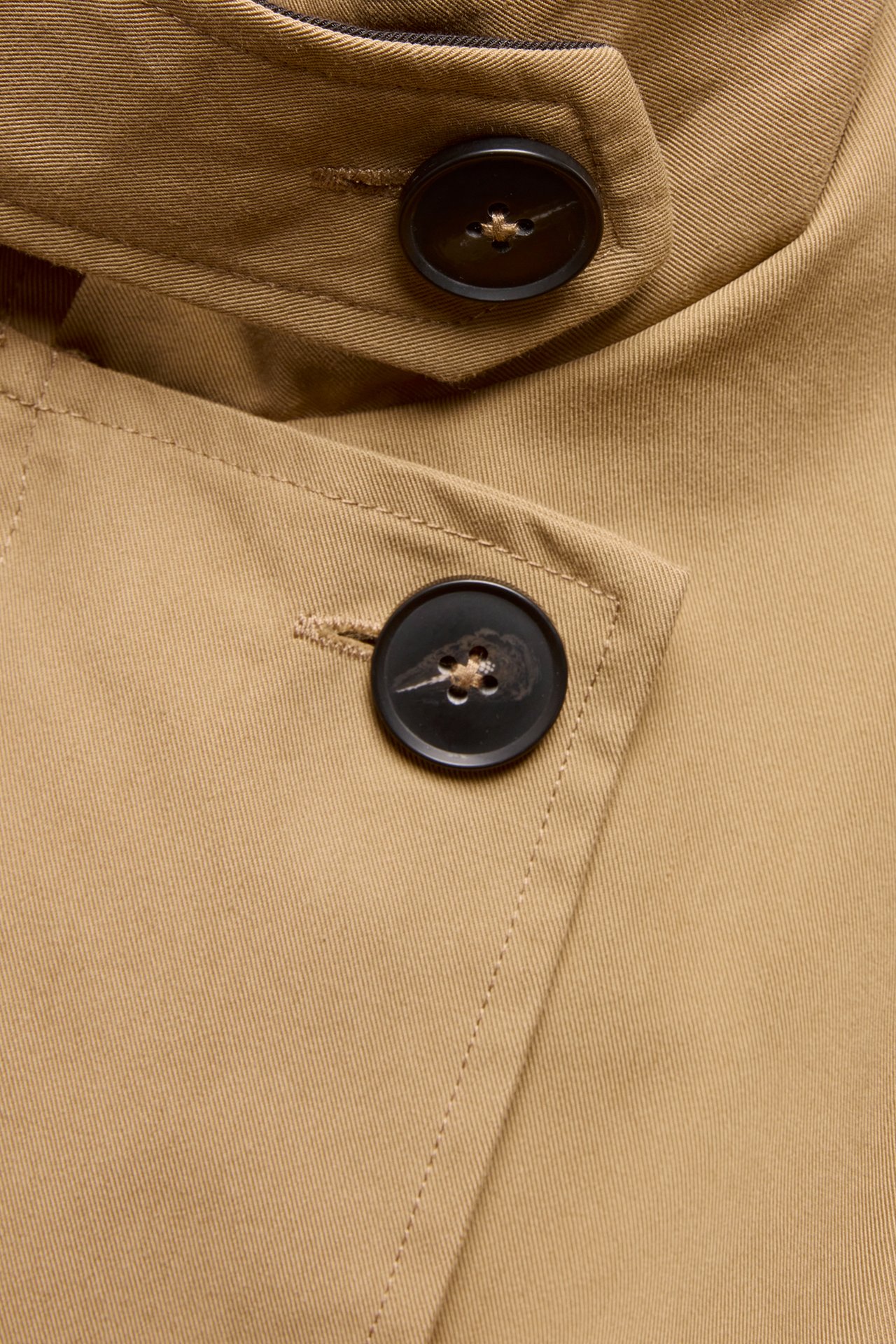 Nærbilde av en beige trenchcoat i bomullsblanding med mørke knapper og sømmer.