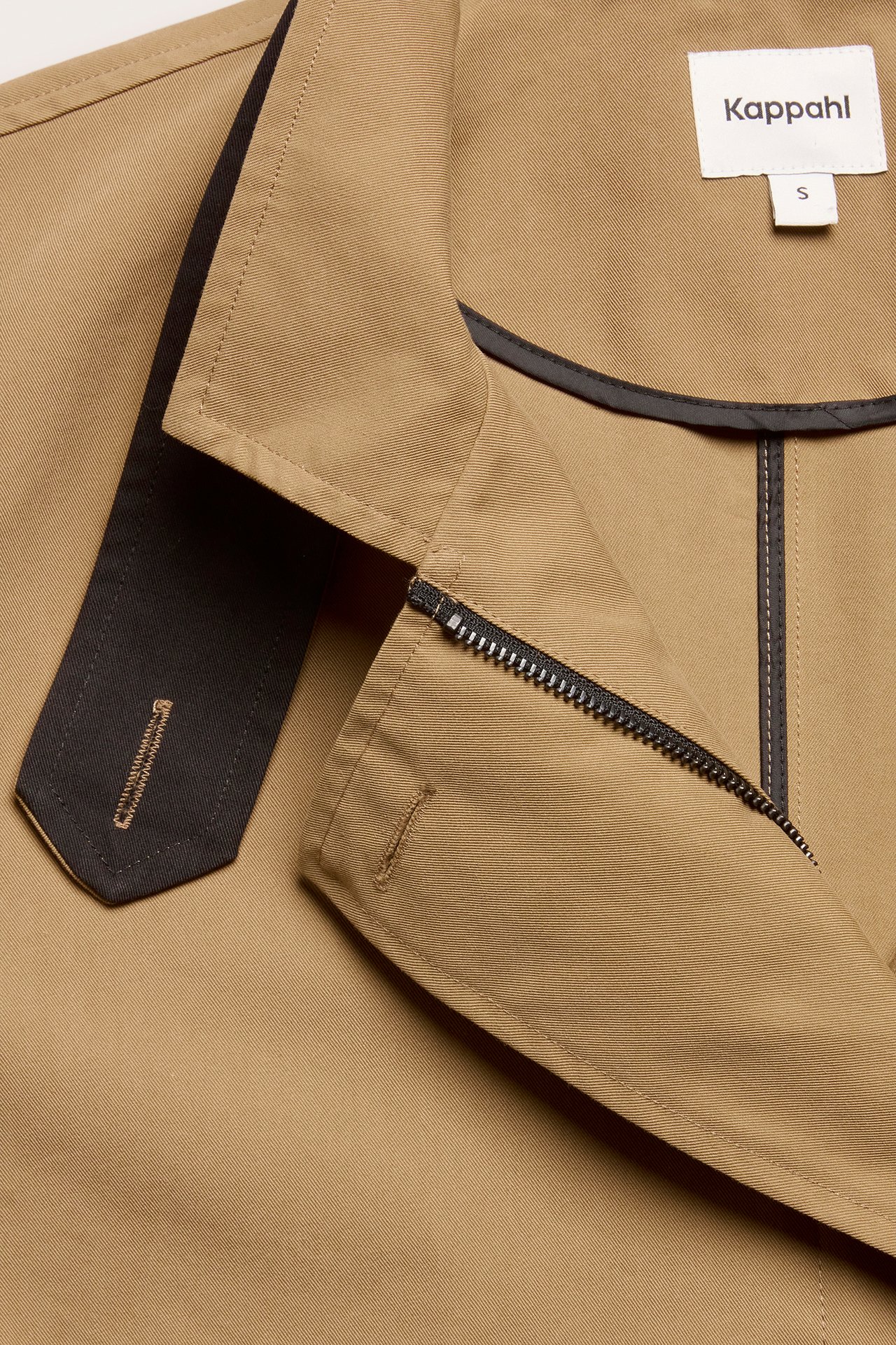 Nærbilde av en beige trenchcoat med svart glidelås, kragekant og knapphullsrem.