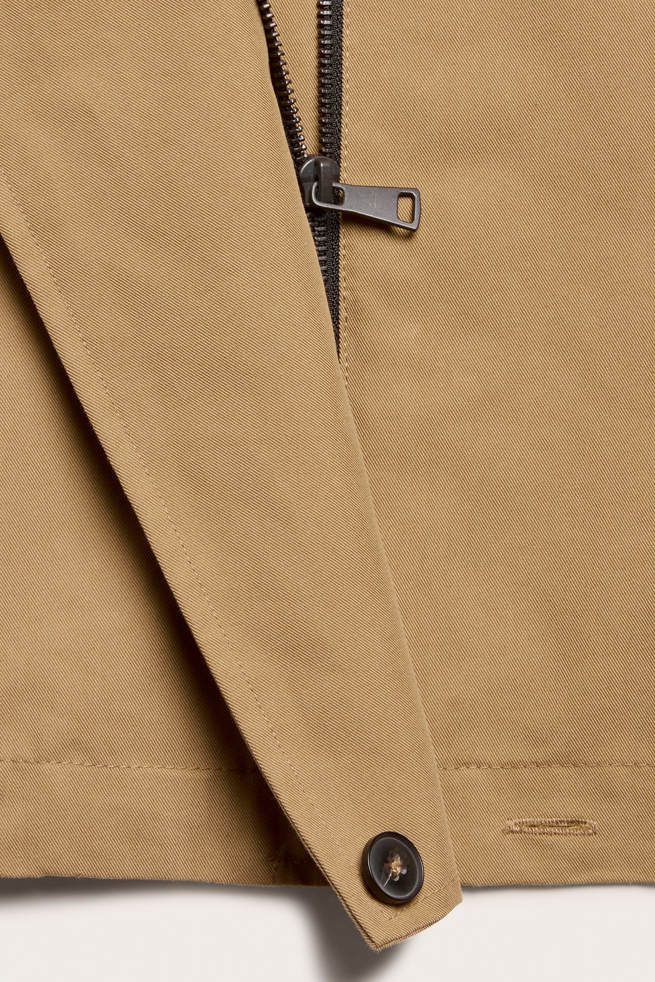 Nærbilde av en beige trenchcoat i bomullsblanding, med svart glidelås og mørk knapp.