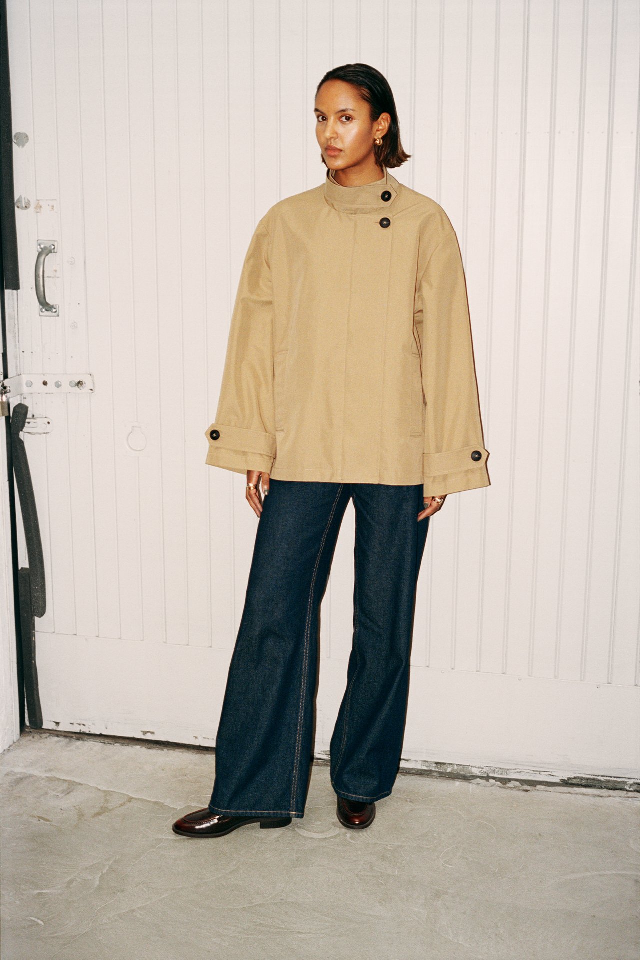 Forfra: kvinne i beige kort trenchcoat med svarte knapper, mørkeblå jeans og brune sko.