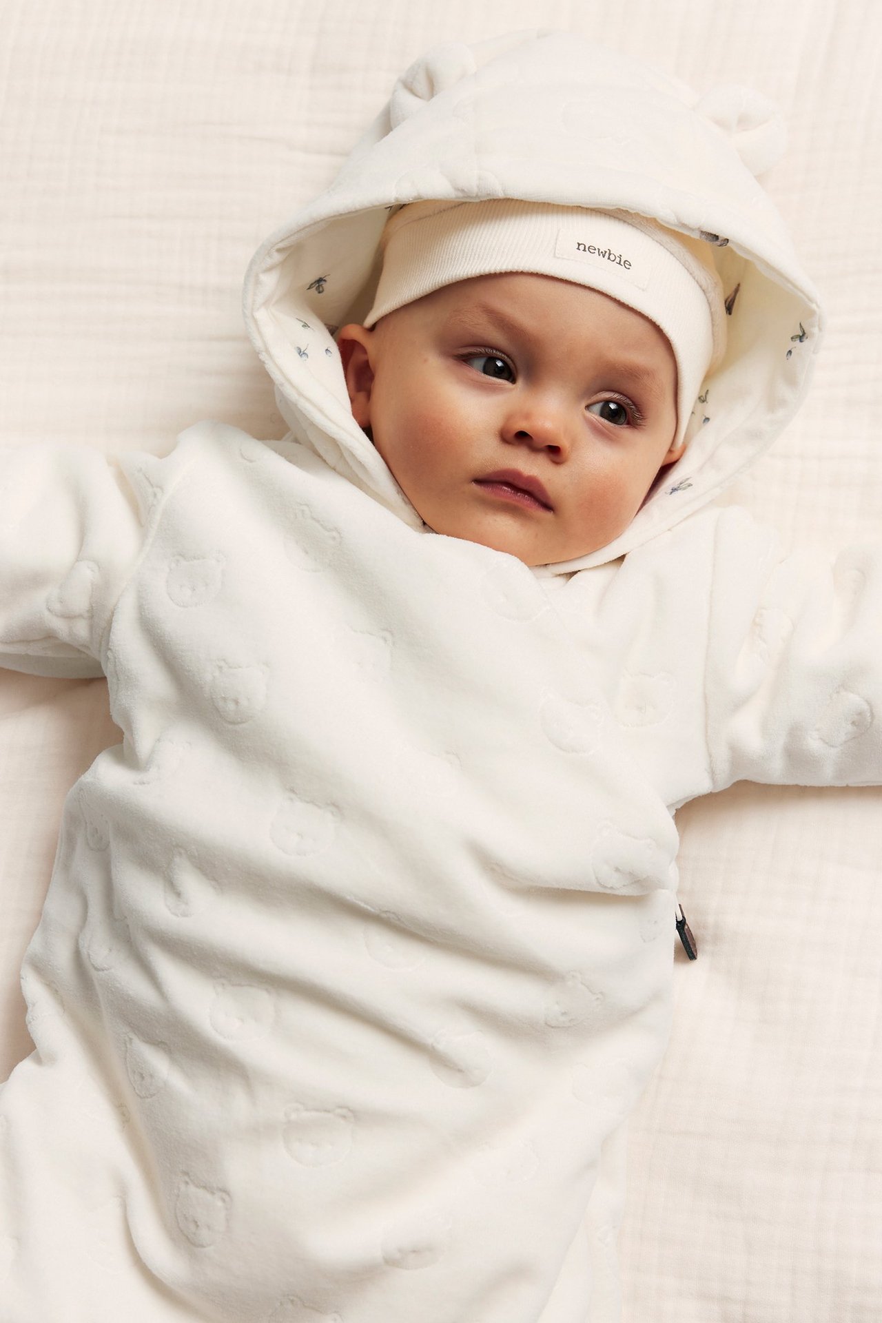 Närbild på baby i offwhite velouroverall med präglade björnar, huva med öron och mössa.