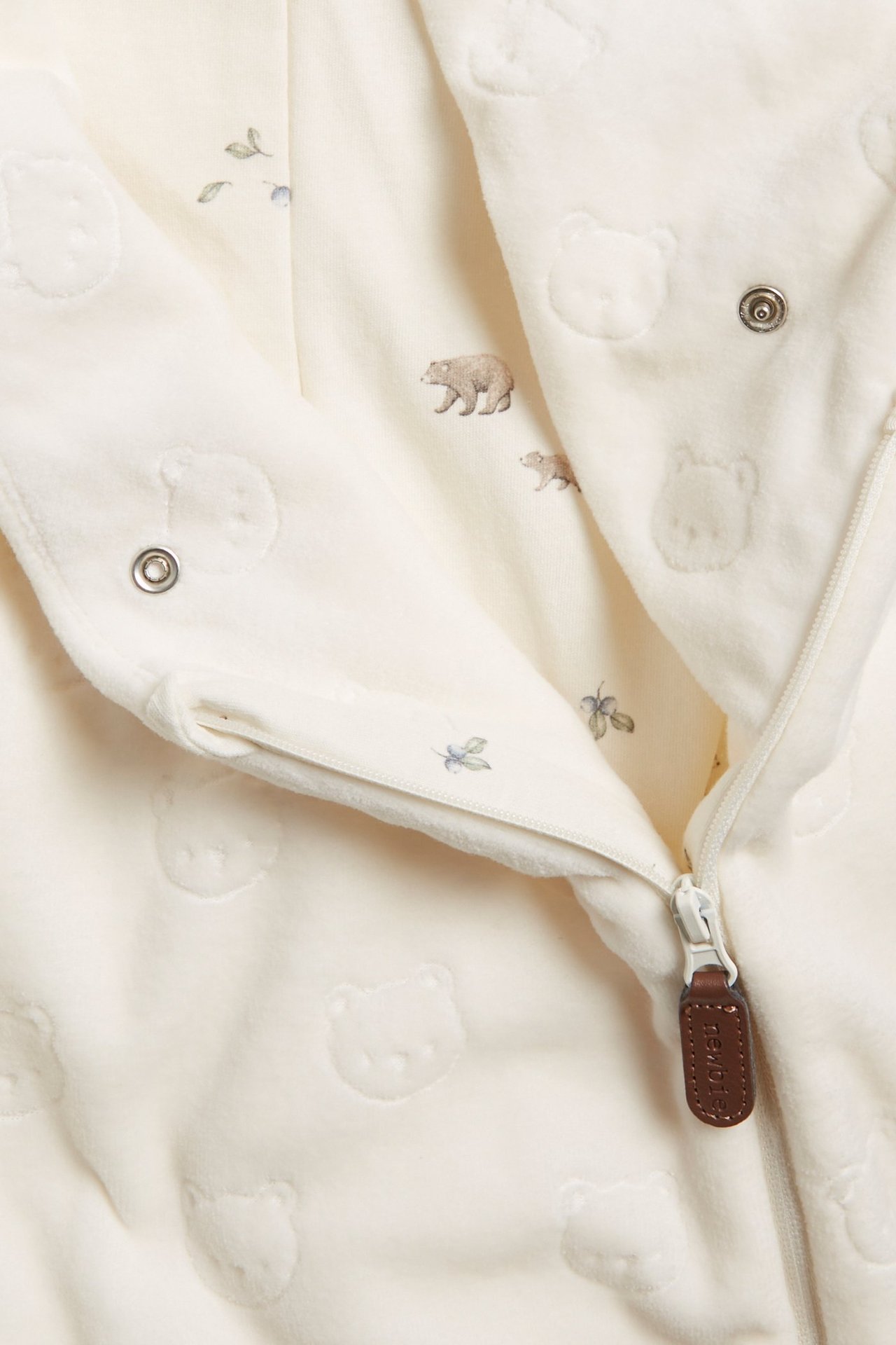 Zbliżenie na ecru welurowy kombinezon niemowlęcy z wytłoczonym wzorem misia i nadrukowaną podszewką, ukośny zamek.