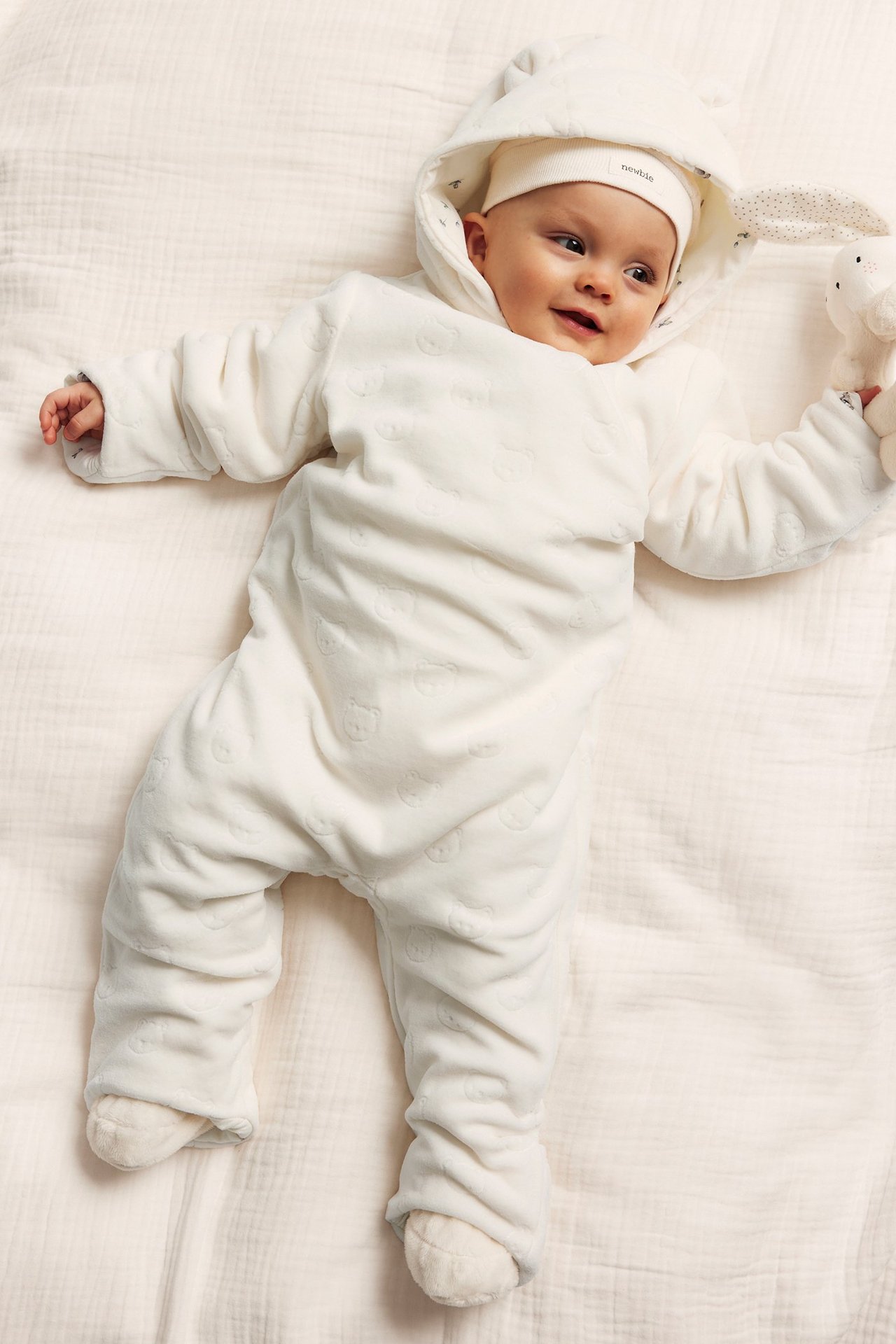 Framifrån: baby i offwhite velouroverall med präglade björnar, huva och håller en vit mjukisleksak.