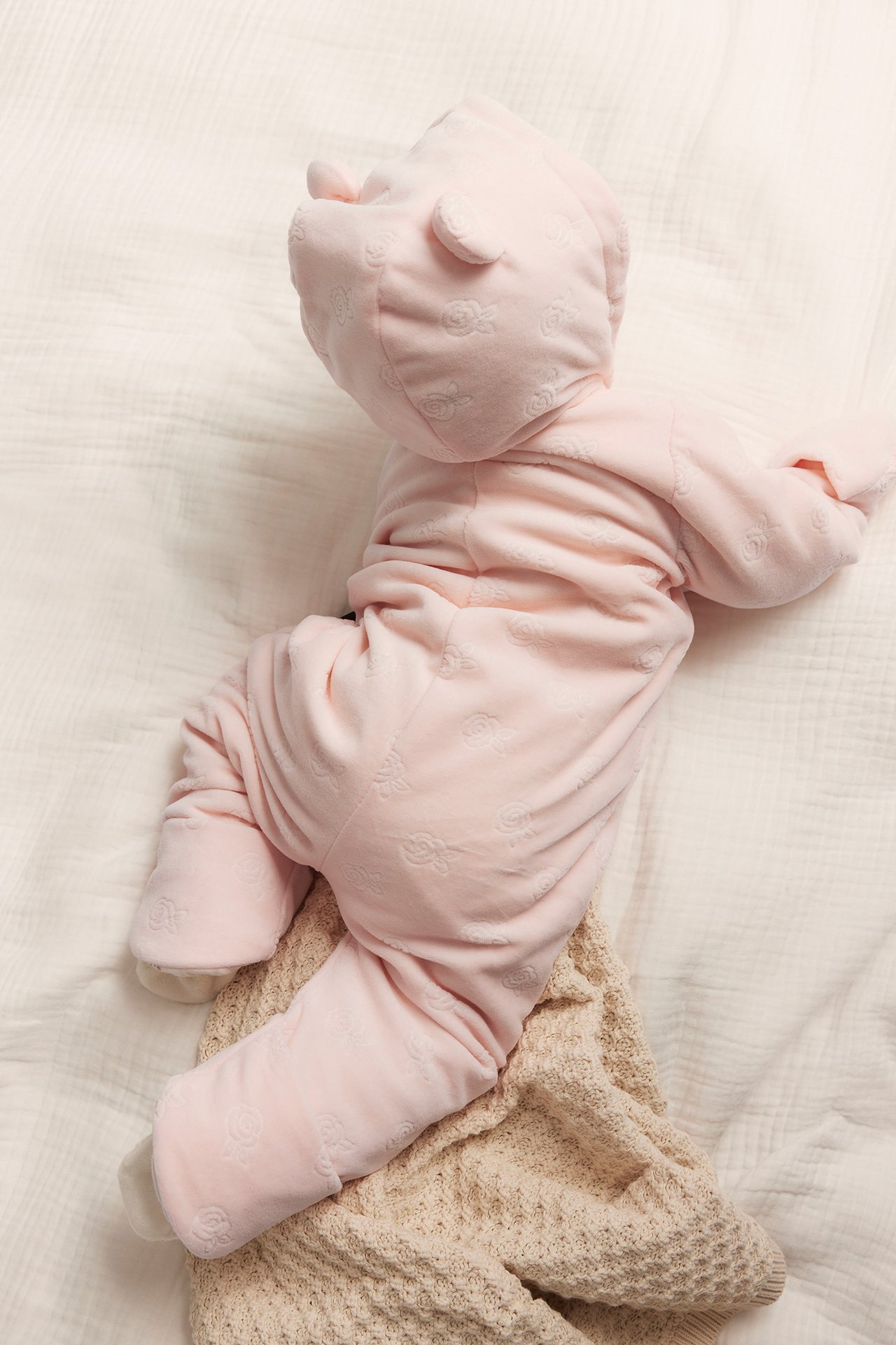 Uppifrån, baby i rosa velouroverall med präglat mönster och huva med öron, liggande på filt.