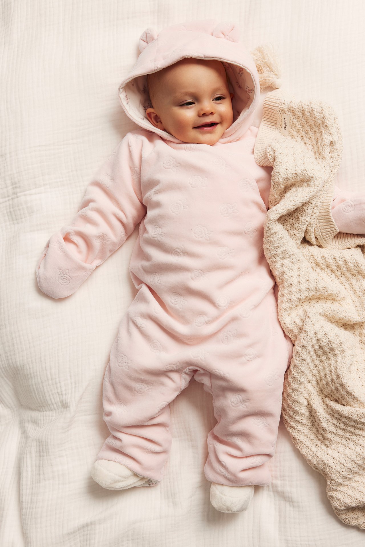 Baby i rosa velouroverall med präglat rosmönster och öronhuva, bredvid en beige stickad filt.