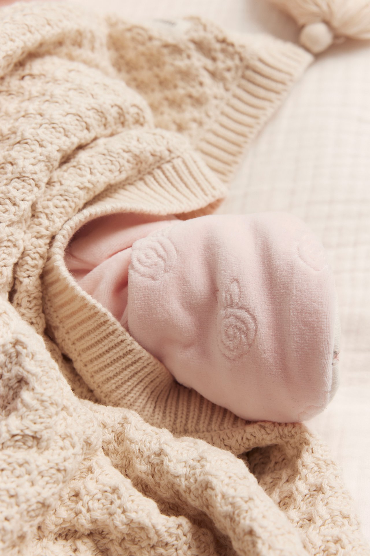 Närbild på babyfot i rosa velouroverall med präglat mönster, under en beige stickad filt.