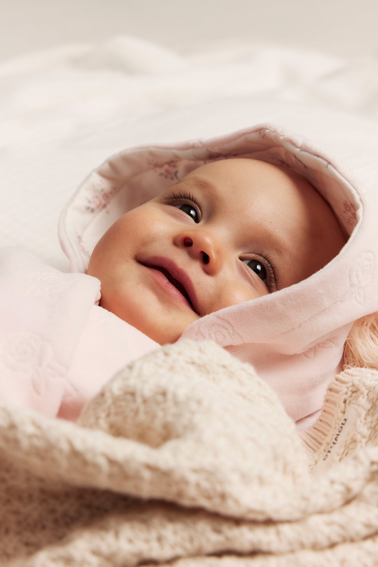 Närbild på baby i rosa velouroverall med präglat mönster och mönstrad huva, under en stickad filt.