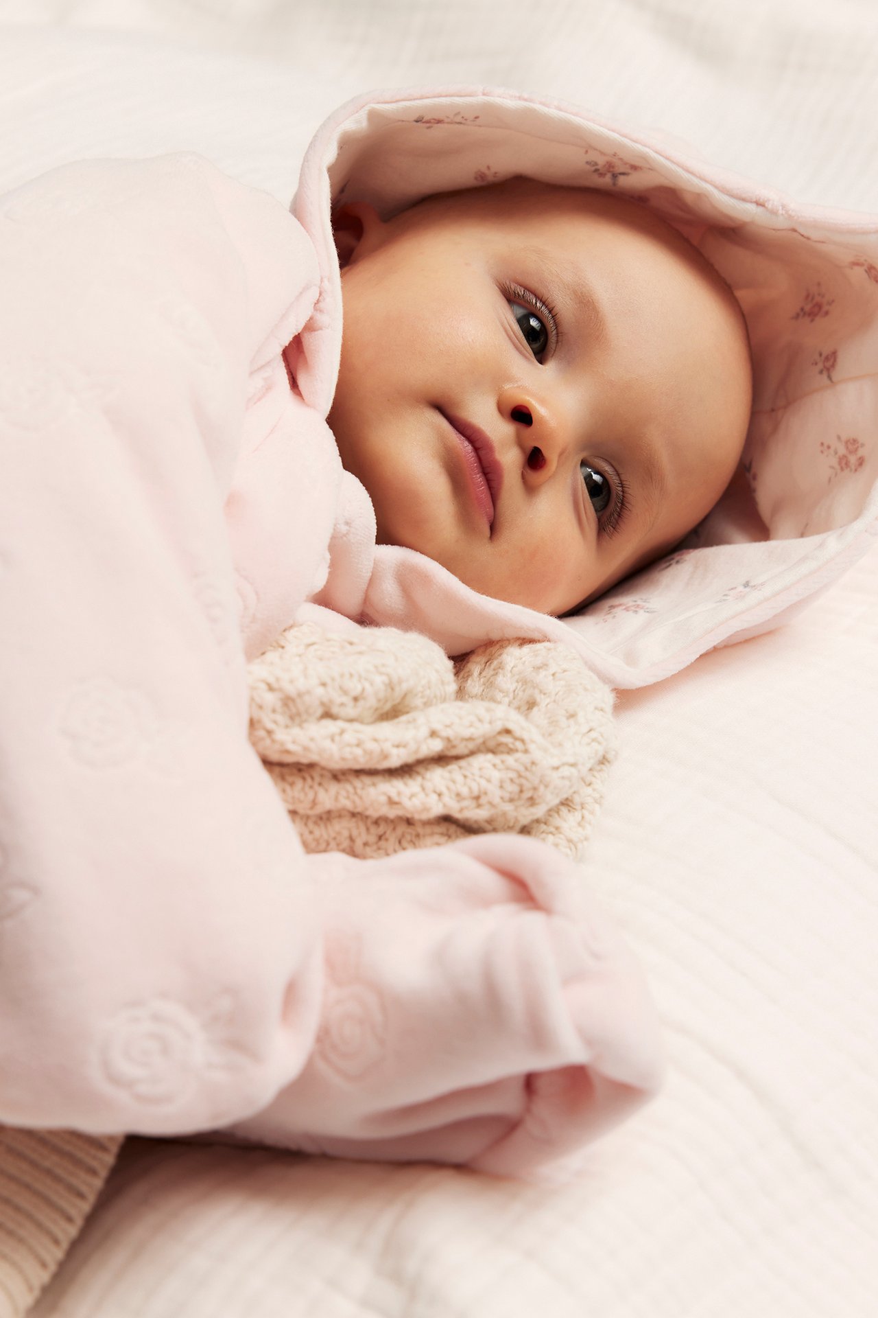 Närbild på en baby i rosa velouroverall med präglat mönster och blommig huva, bredvid en beige filt.