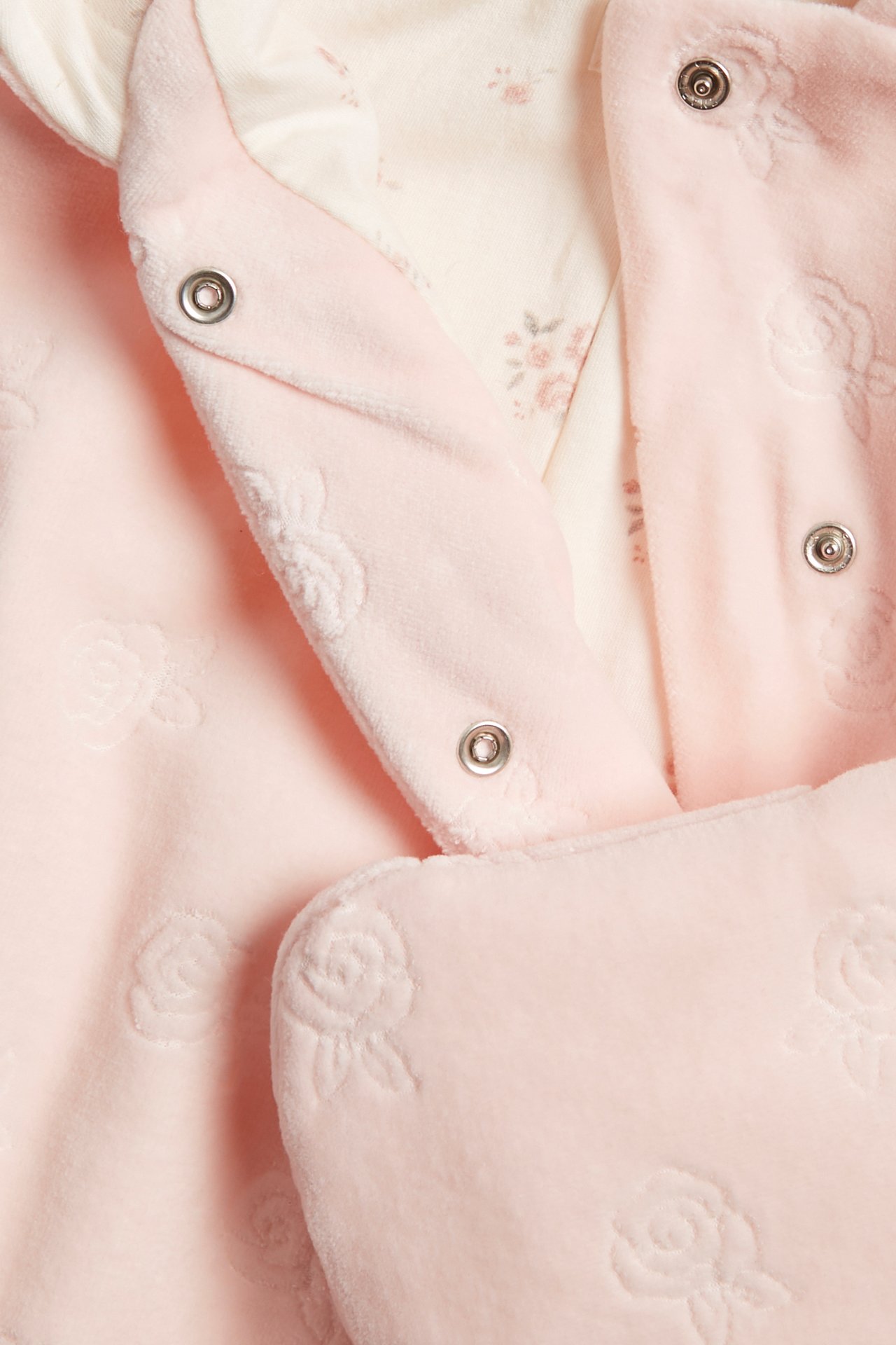Närbild på en rosa velouroverall för baby med präglade rosor, tryckknappar och blommigt foder.