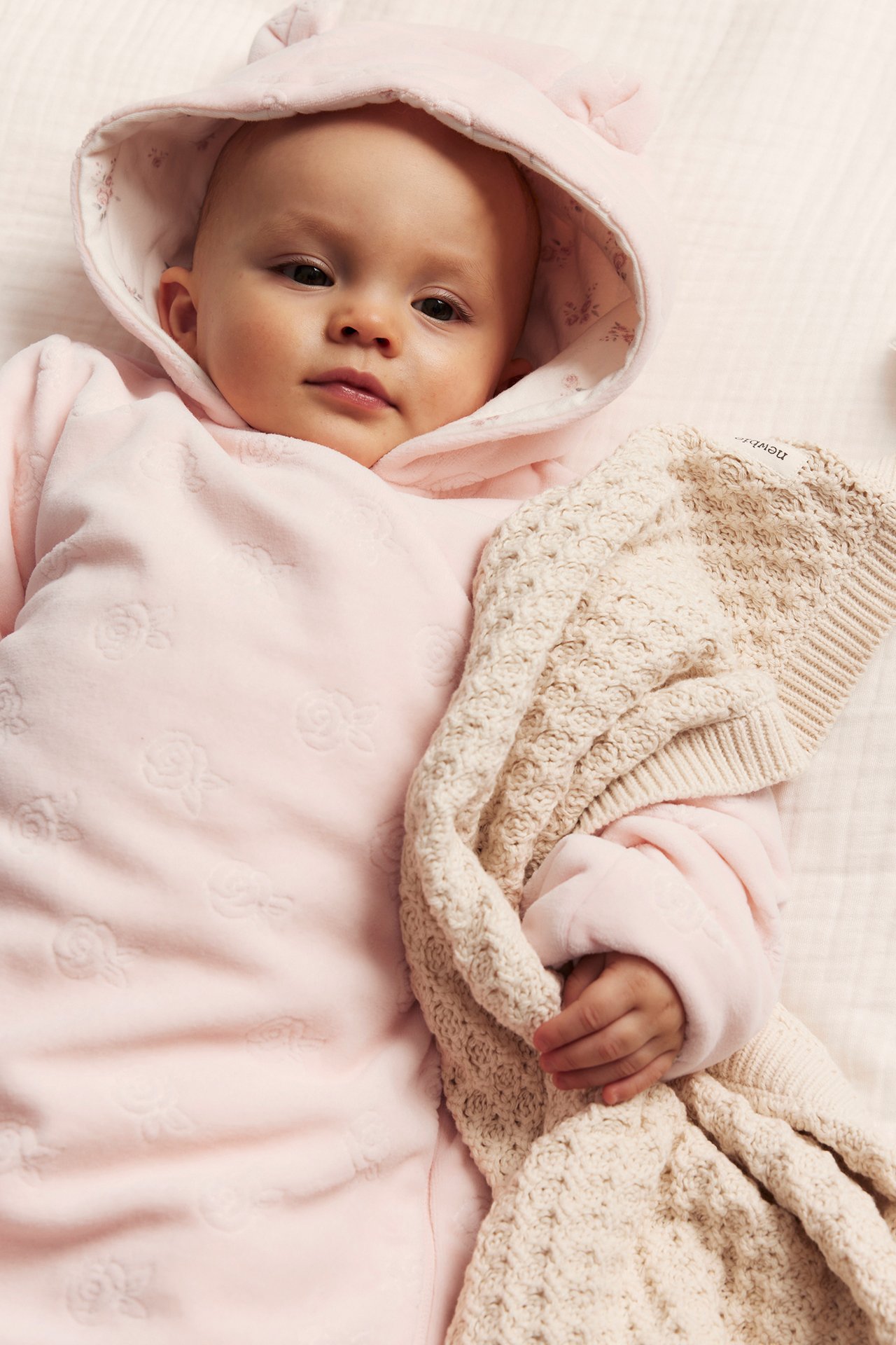 Ovanifrån: Baby i rosa velouroverall med präglade rosor, öronhuva, håller en beige stickad filt.