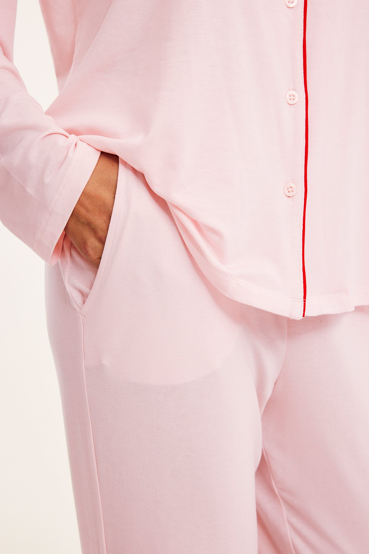 Lähikuva: Nainen vaaleanpunaisessa lyocell-pyjamassa, napitettu paita punaisella tereellä, housut käsi taskussa.