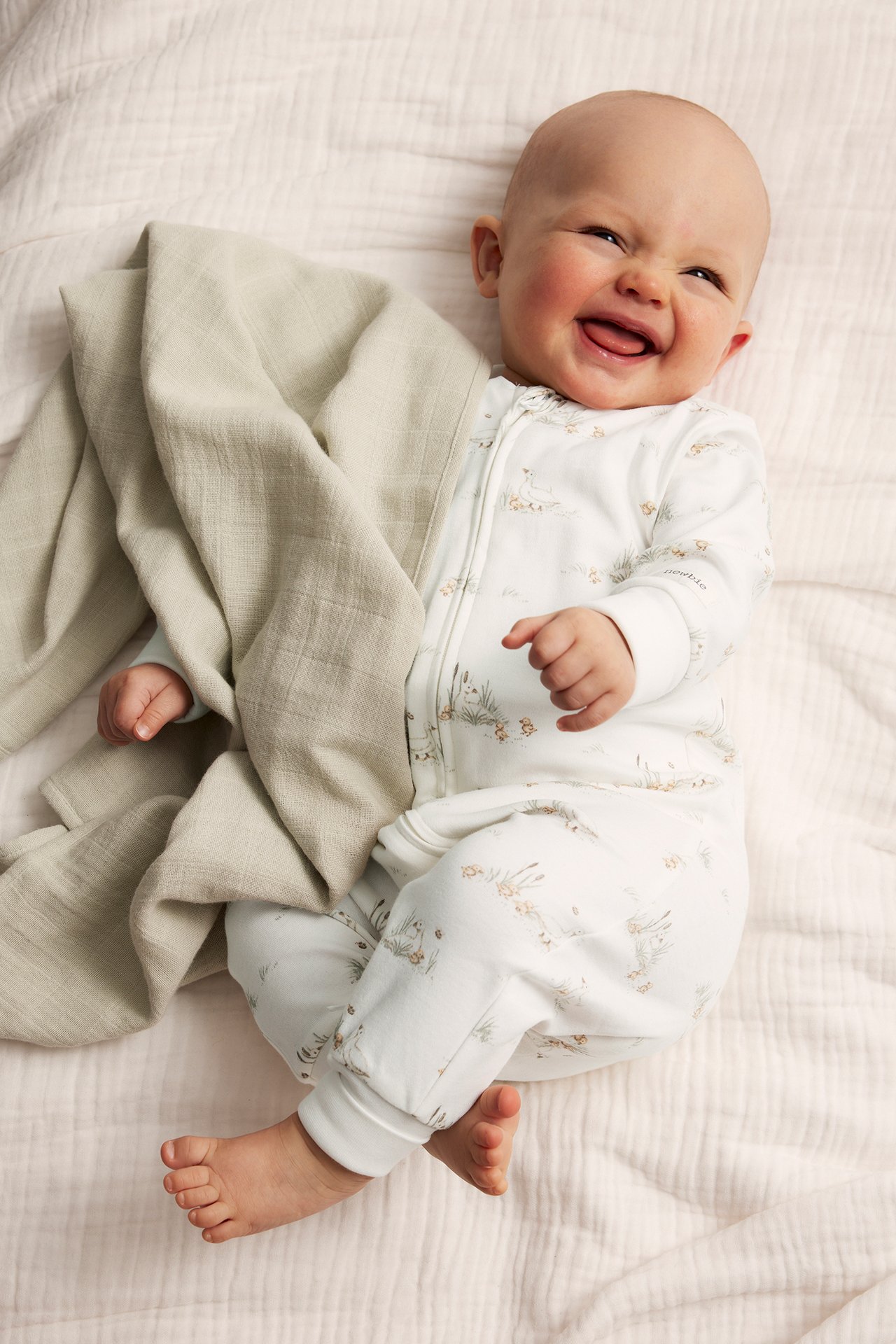 Hymyilevä vauva offwhite-puuvillapyjamassa, jossa ankka-kuvio ja vetoketju, vaaleanvihreän peiton vieressä.