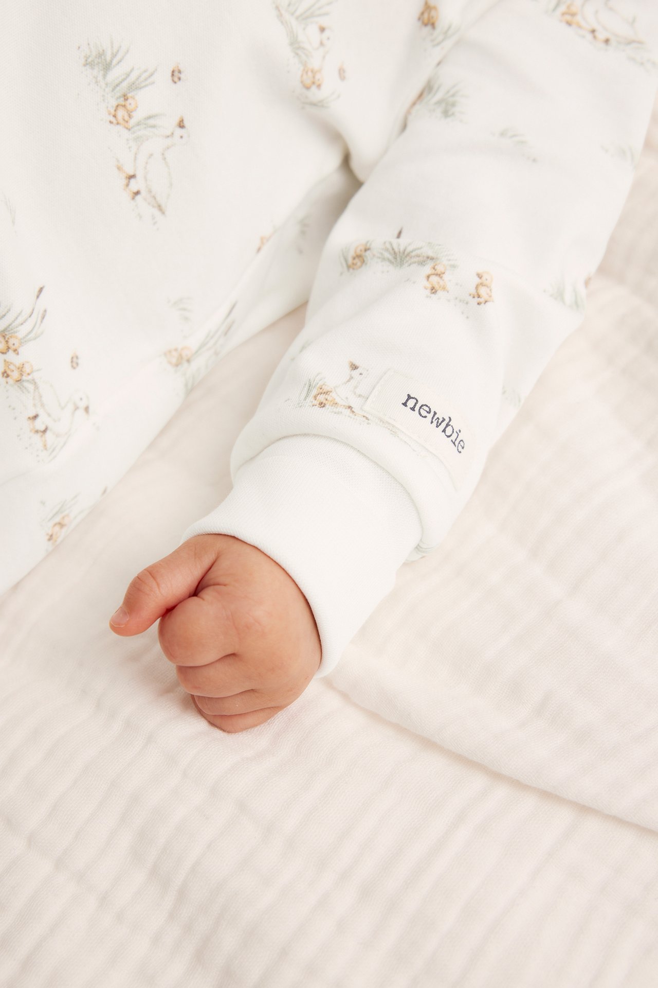 Lähikuva vauvan käsivarresta offwhite-puuvillapyjamassa, jossa ankka-kuvio ja 'newbie'-merkki.