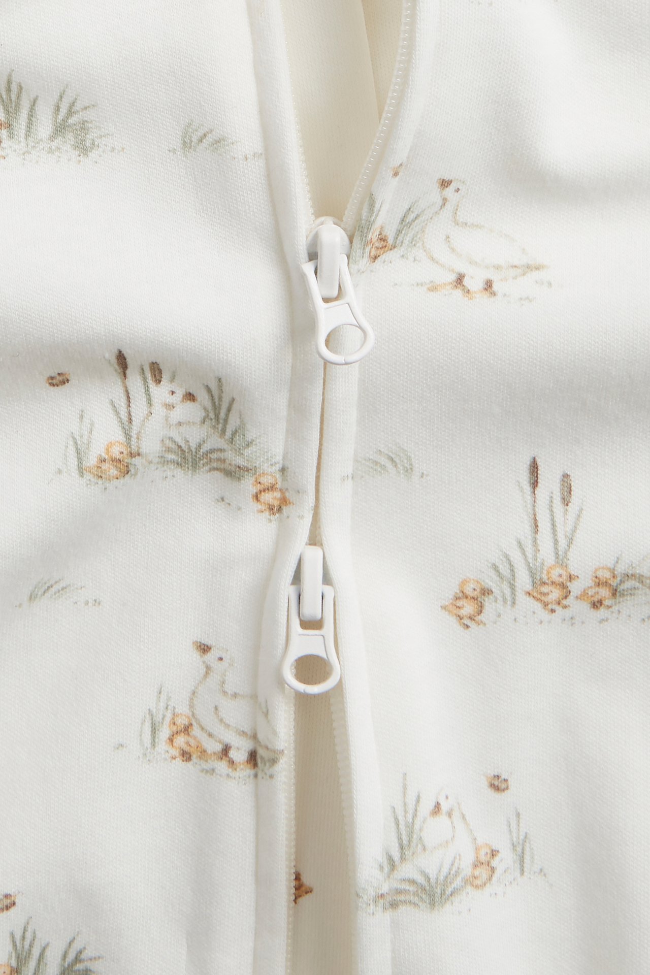 Lähikuva luonnonvalkoisesta puuvillapyjamasta, jossa ankka-kuvio ja valkoinen kaksisuuntainen vetoketju.