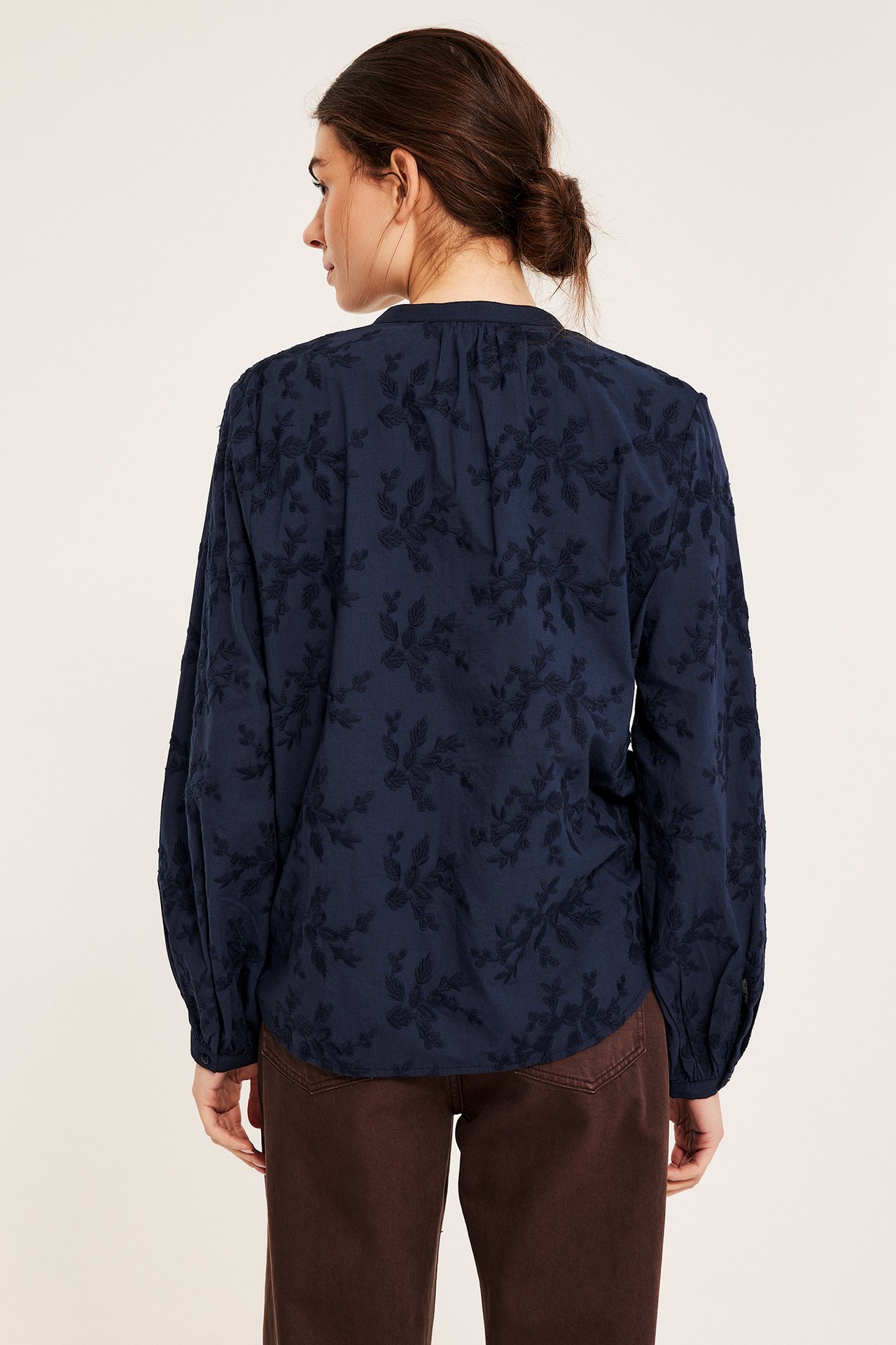 Widok z tyłu kobiety w granatowej bawełnianej bluzce z haftowanymi kwiatami, noszonej z brązowymi spodniami.