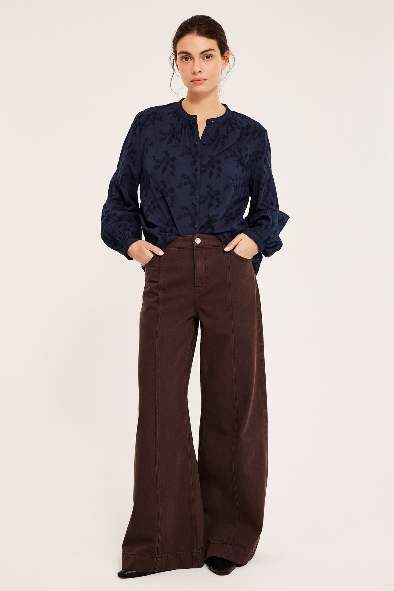 Widok z przodu kobiety w granatowej bawełnianej bluzce z haftowanymi kwiatami i ciemnobrązowych szerokich spodniach.