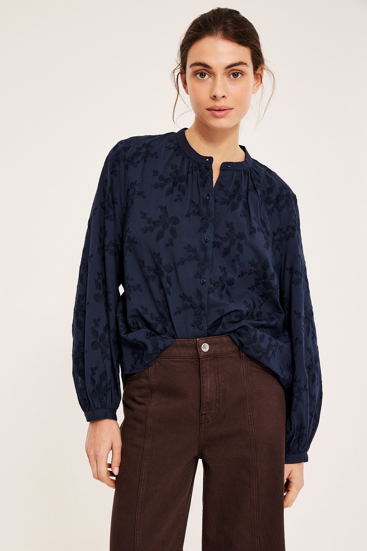 Widok z przodu kobiety w granatowej bawełnianej bluzce z haftem kwiatowym, guzikami i brązowych spodniach.