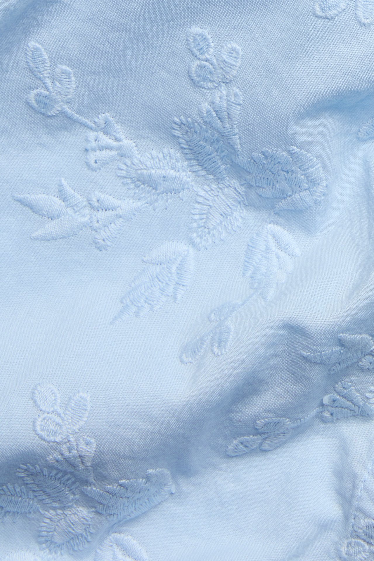Lähikuva vaaleansinisestä puuvillapuserosta, jossa on brodeerattuja kukkakuvioita.