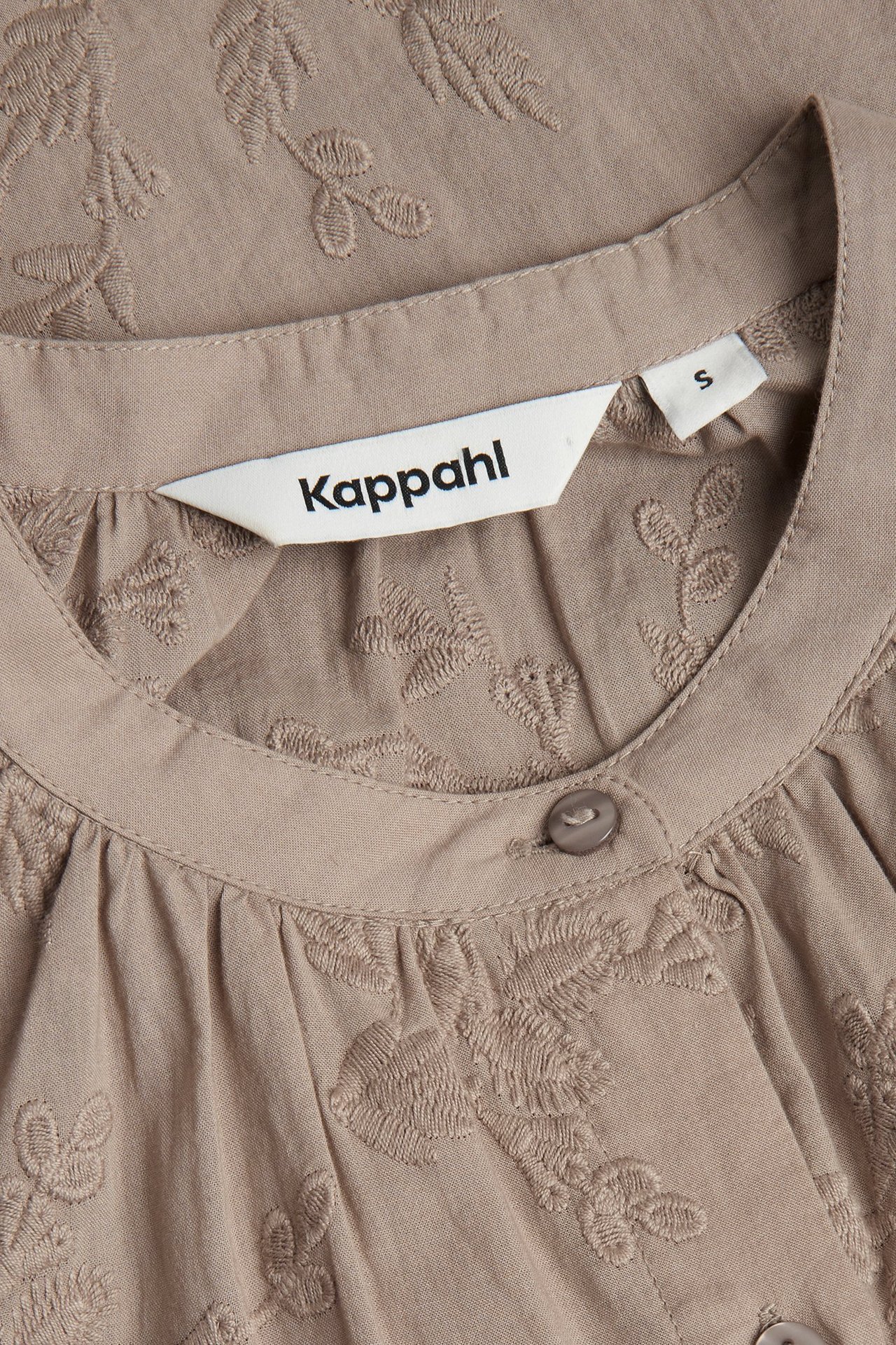 Zbliżenie na ciemnobeżową bawełnianą bluzkę z haftowanymi kwiatowymi wzorami i guzikami przy dekolcie.