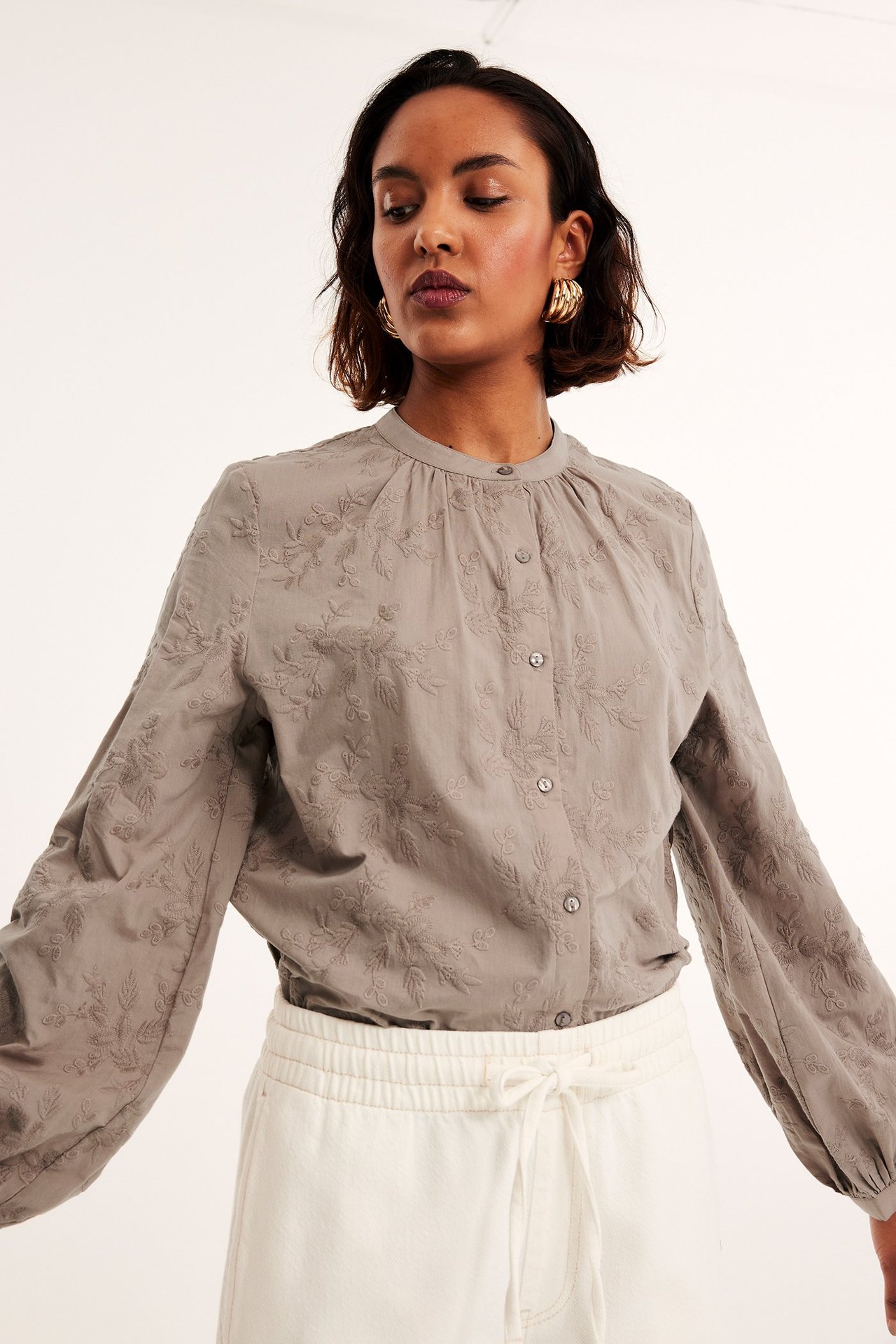 Widok z przodu kobiety w ciemnobeżowej bawełnianej bluzce z haftowanymi kwiatami, guzikami i białymi spodniami.