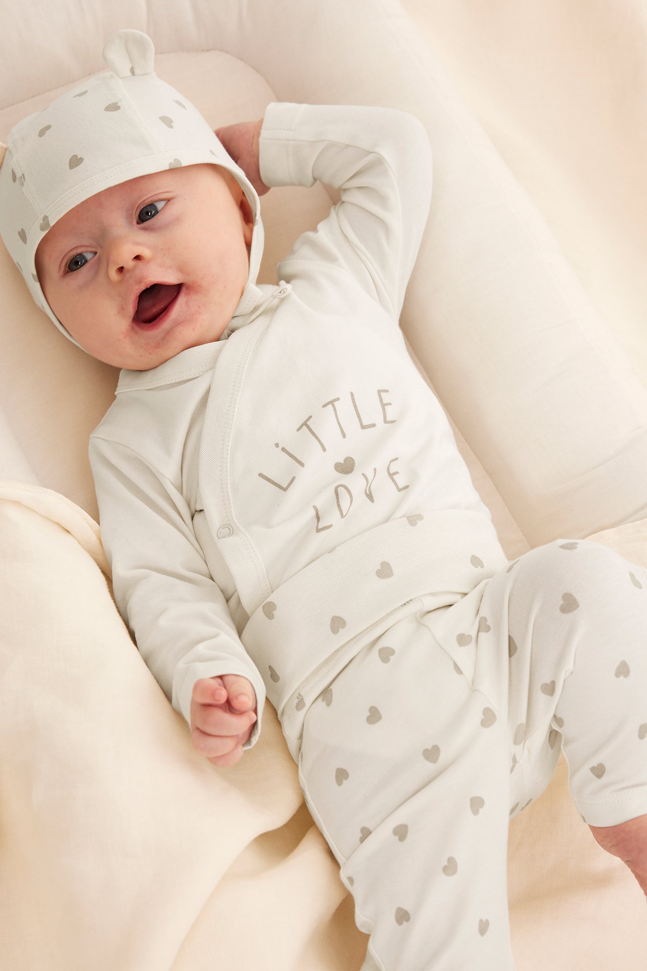 Högvinkel framifrån av baby i offwhite mönstrad omlottbody, matchande byxor och mössa med öron.