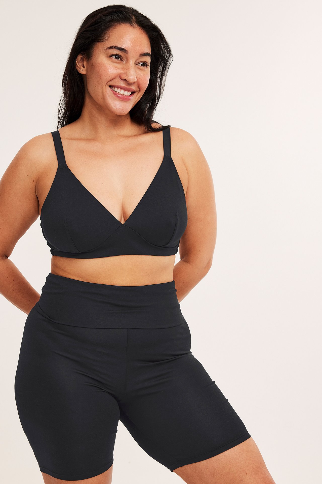 Frontvy av en leende kvinna i svarta lyocellshorts med brett midjeband och en svart bralette.