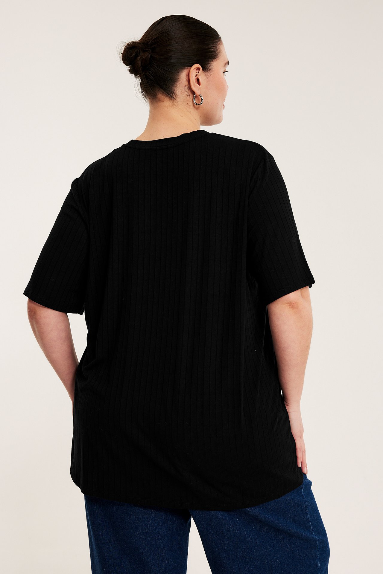 Lång svart ribbad t-shirt på en kvinna, med mörkblå jeans och silverörhängen, bakifrån.
