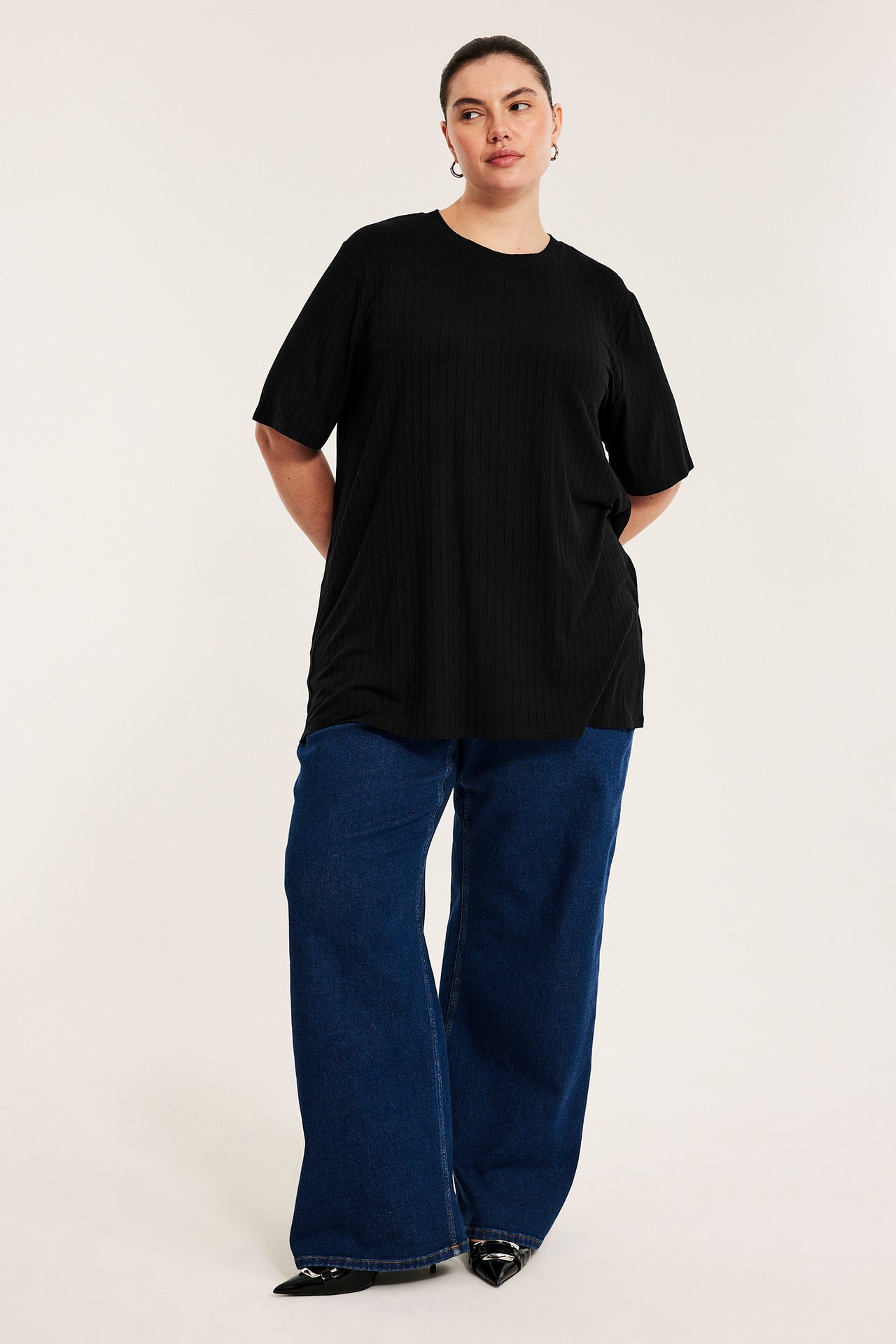 Framifrån, kvinna i lång svart ribbad t-shirt, vida mörkblå jeans och svarta spetsiga skor.