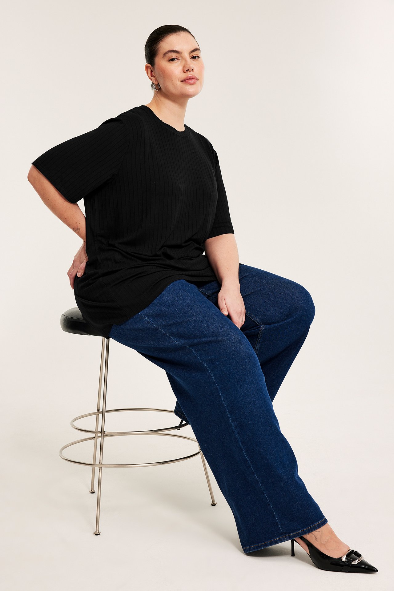 Frontvy av en kvinna i en lång svart ribbad t-shirt, mörkblå vida jeans och svarta högklackade skor.