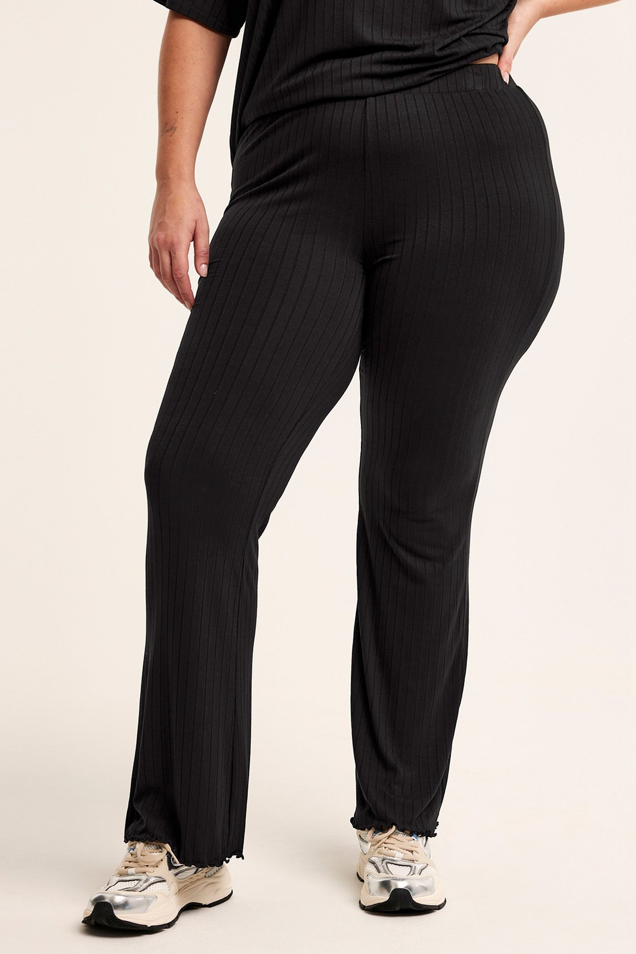 Framifrån: Kvinna i svarta ribbade utsvängda jerseybyxor med volangfåll, matchande topp och ljusa sneakers.