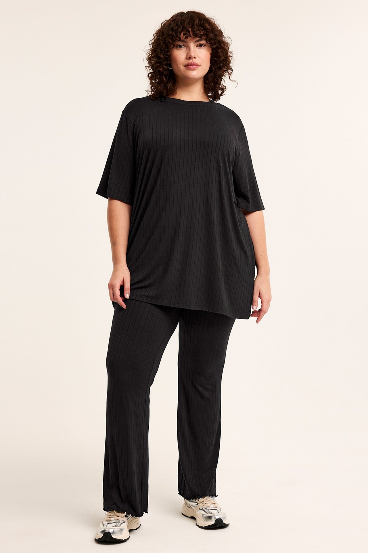 Frontvy av kvinna i svarta ribbade utsvängda jerseybyxor med salladskant och matchande oversized t-shirt.