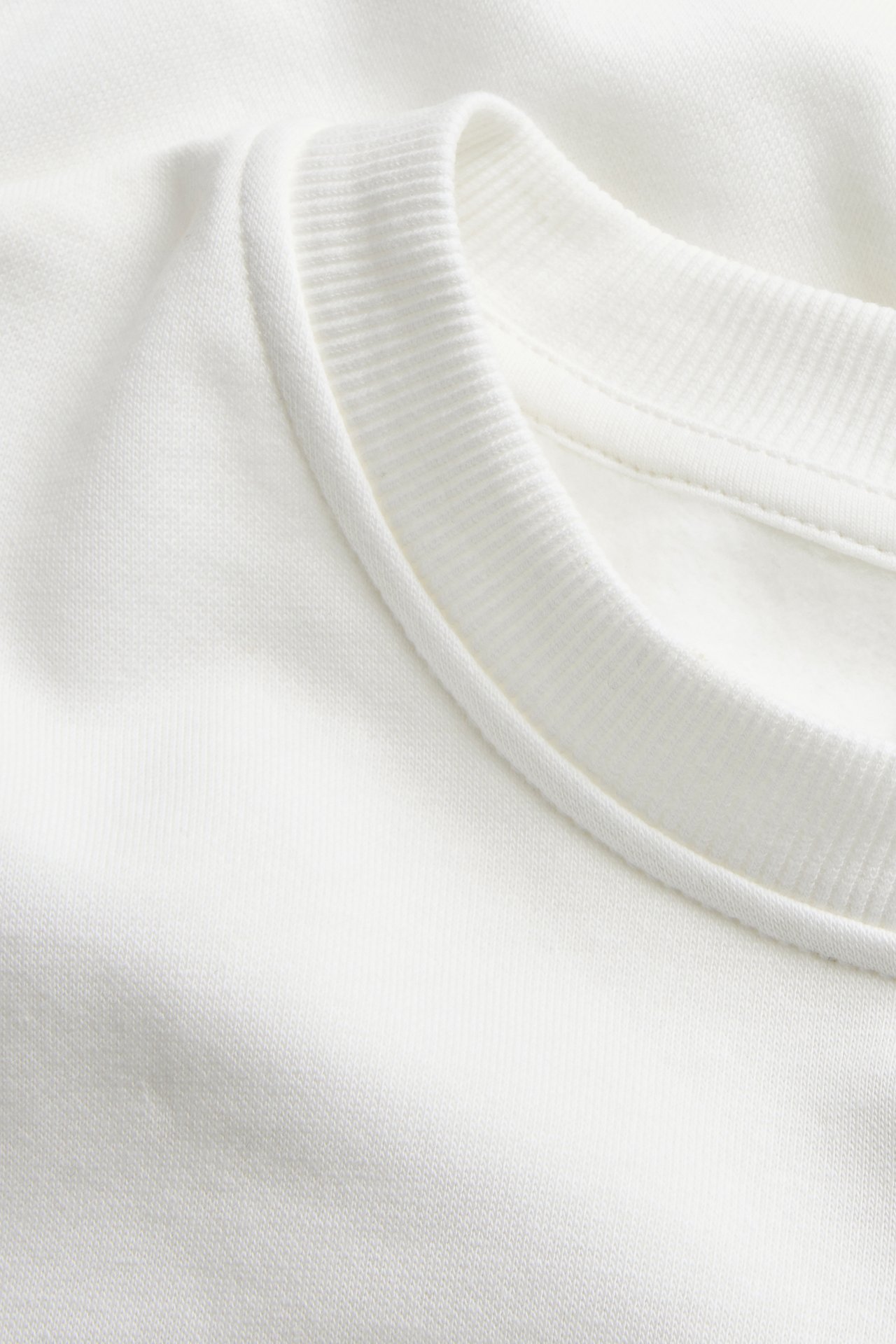 Närbild av en offwhite enfärgad sweatshirt med ribbad rundhals i bomullsblandning.
