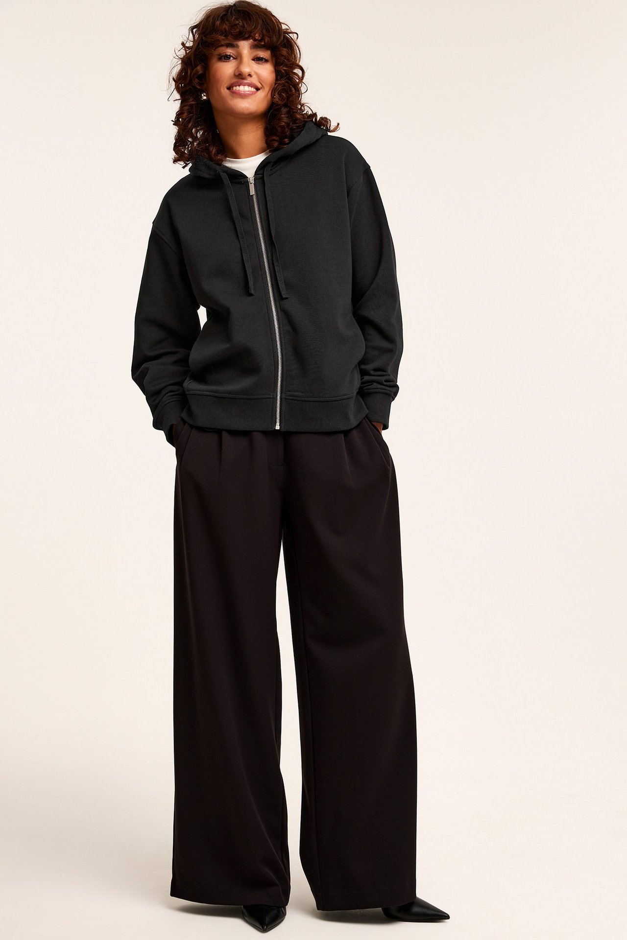 Widok z przodu: kobieta w czarnej bluzie z kapturem, białym t-shircie, czarnych szerokich spodniach i butach.