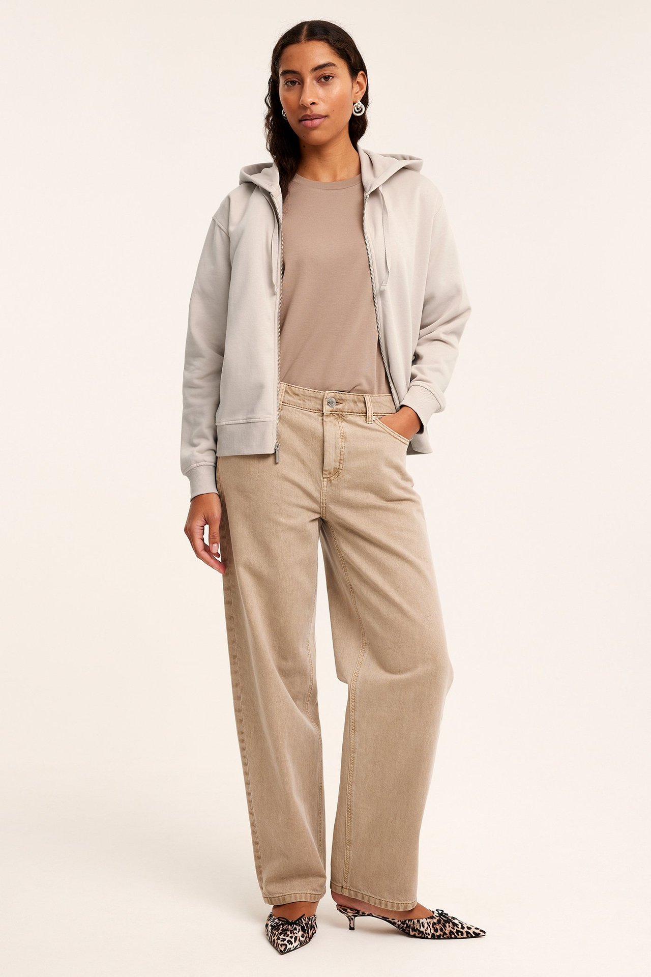 Widok z przodu: Kobieta w otwartej beżowej bluzie z zamkiem, beżowym t-shircie, jasnobrązowych jeansach i mulesach w panterkę.